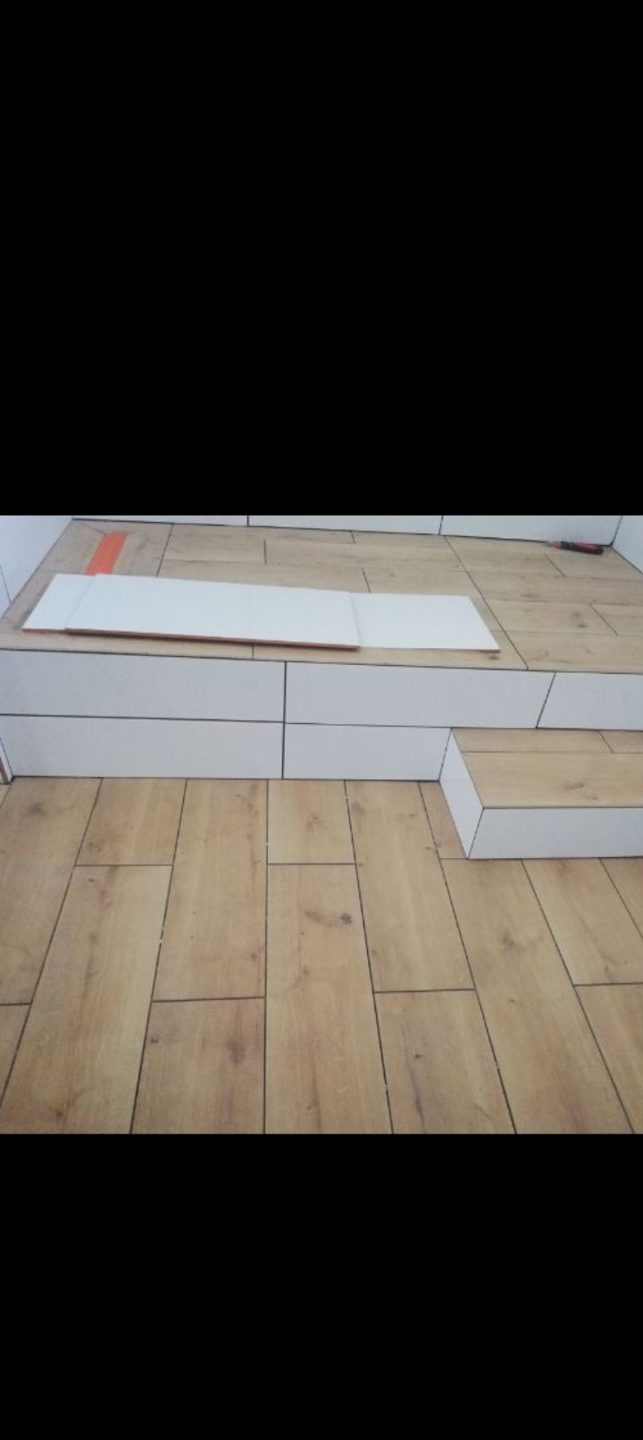 Ułożona podłoga z paneli imitujących drewno, z widocznym fragmentem białej obudowy i narzędziem w tle. Układanie paneli podłogowych.