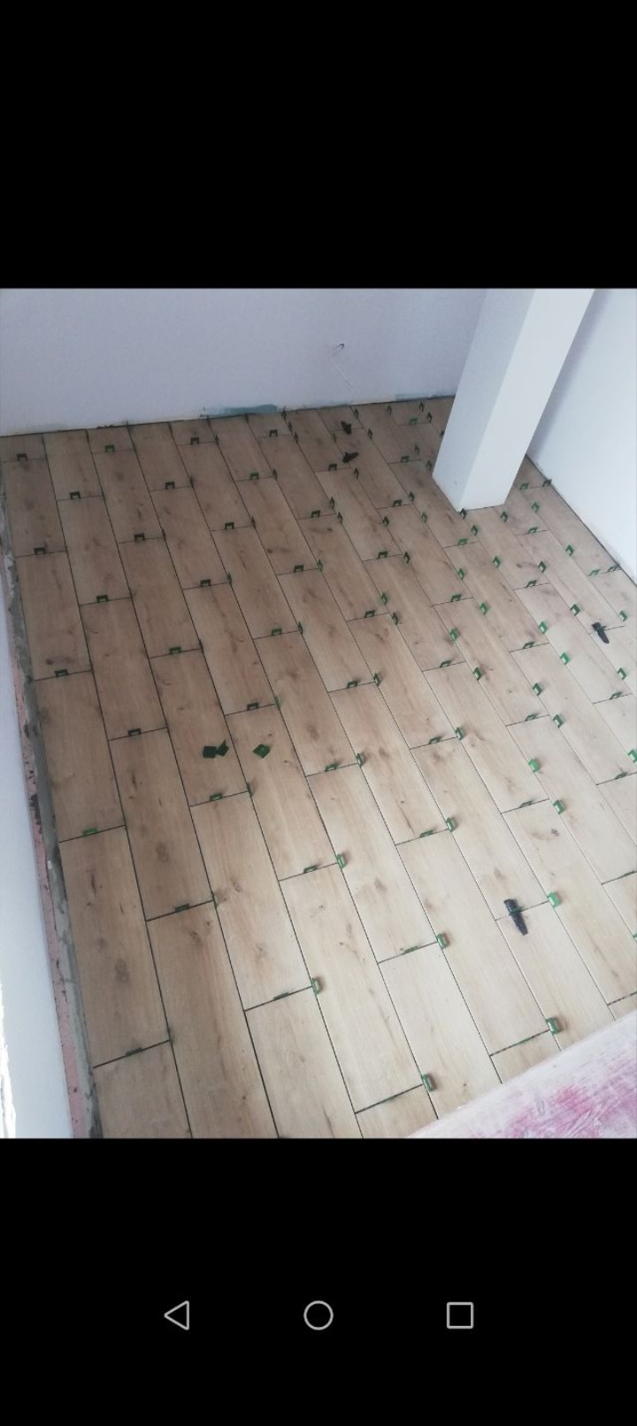 Układanie podłogi z płytek imitujących drewno, z użyciem zielonych klipsów poziomujących. Widoczny fragment ściany i filar. Praca w toku.