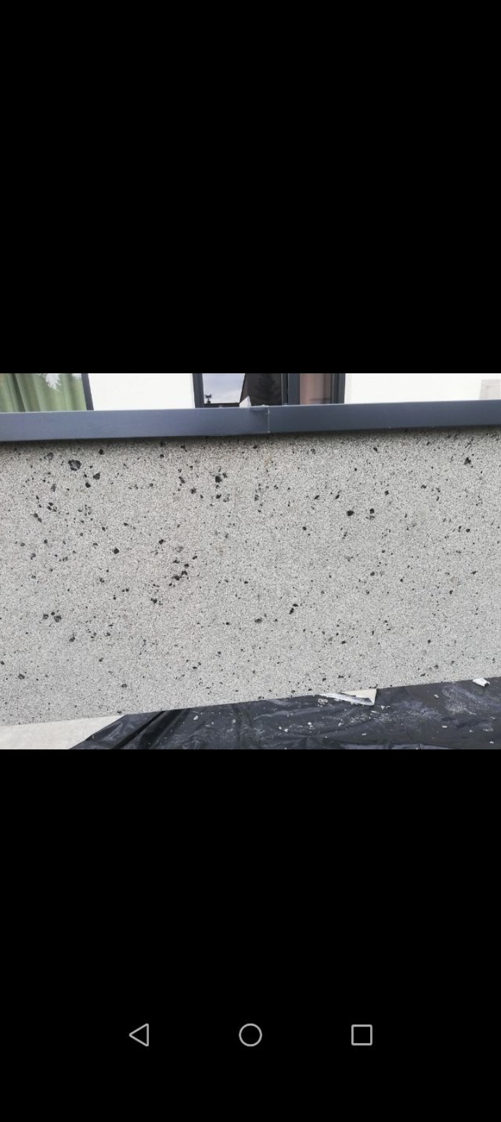 Fragment elewacji z szarą strukturą, zabezpieczonej folią malarską. Widoczne okno i ciemna listwa. Detal wykończenia ściany zewnętrznej.