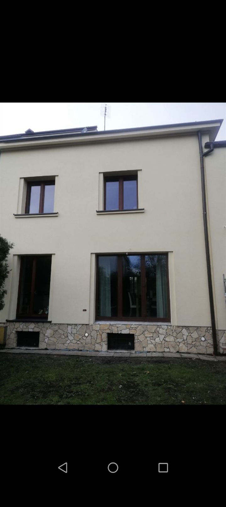 Elewacja domu jednorodzinnego z beżowym tynkiem i kamienną okładziną u podstawy, brązowe okna, widok na ogród i fragment elewacji sąsiedniego budynku.