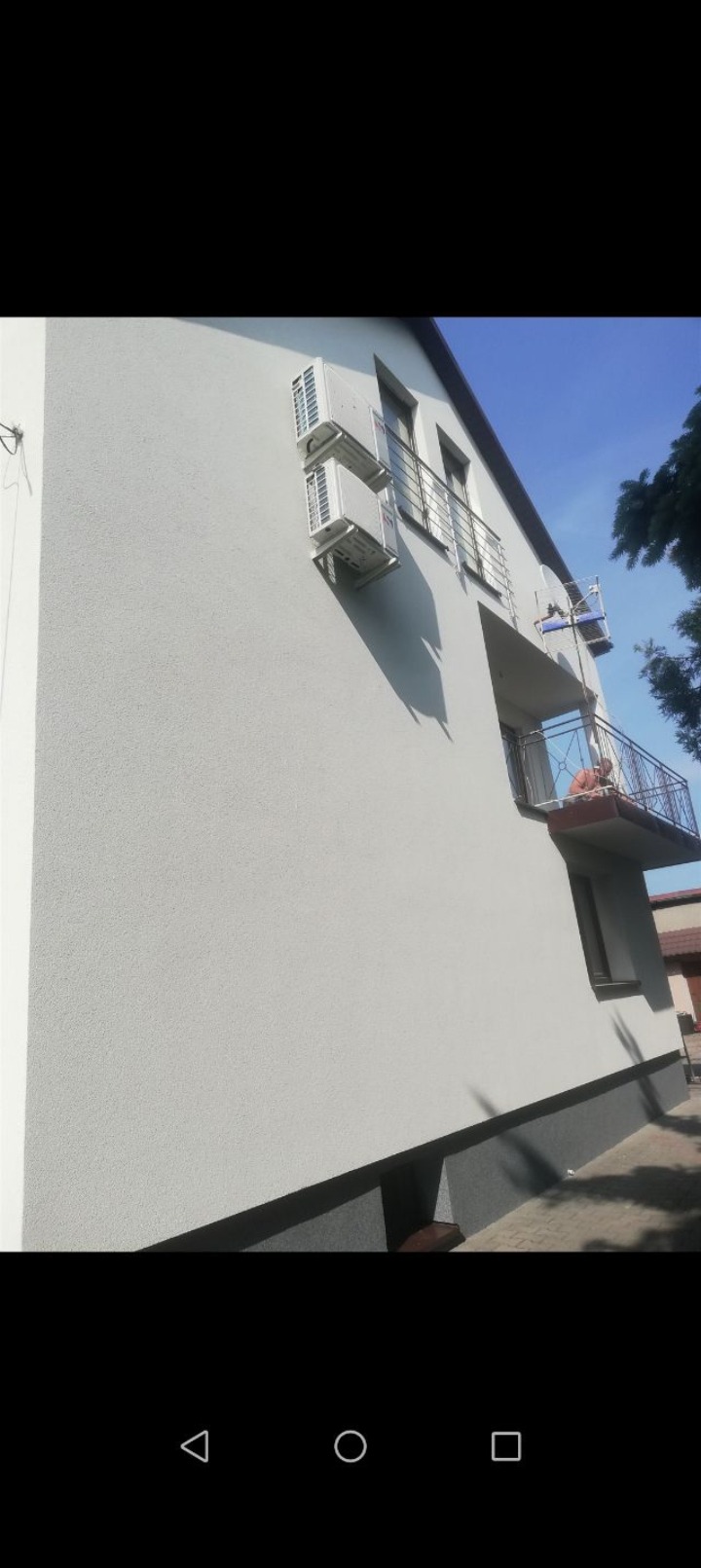 Boczny widok budynku z jasną elewacją, balkonami z metalową balustradą i zamontowanymi klimatyzatorami. Widoczne okna i fragment nieba.