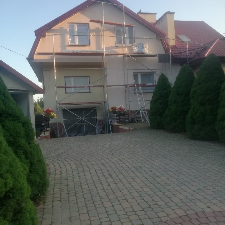 Dom jednorodzinny w trakcie prac elewacyjnych, widoczne rusztowania i zabezpieczone okna. Uwagę zwracają kwiaty w donicach przy wejściu.