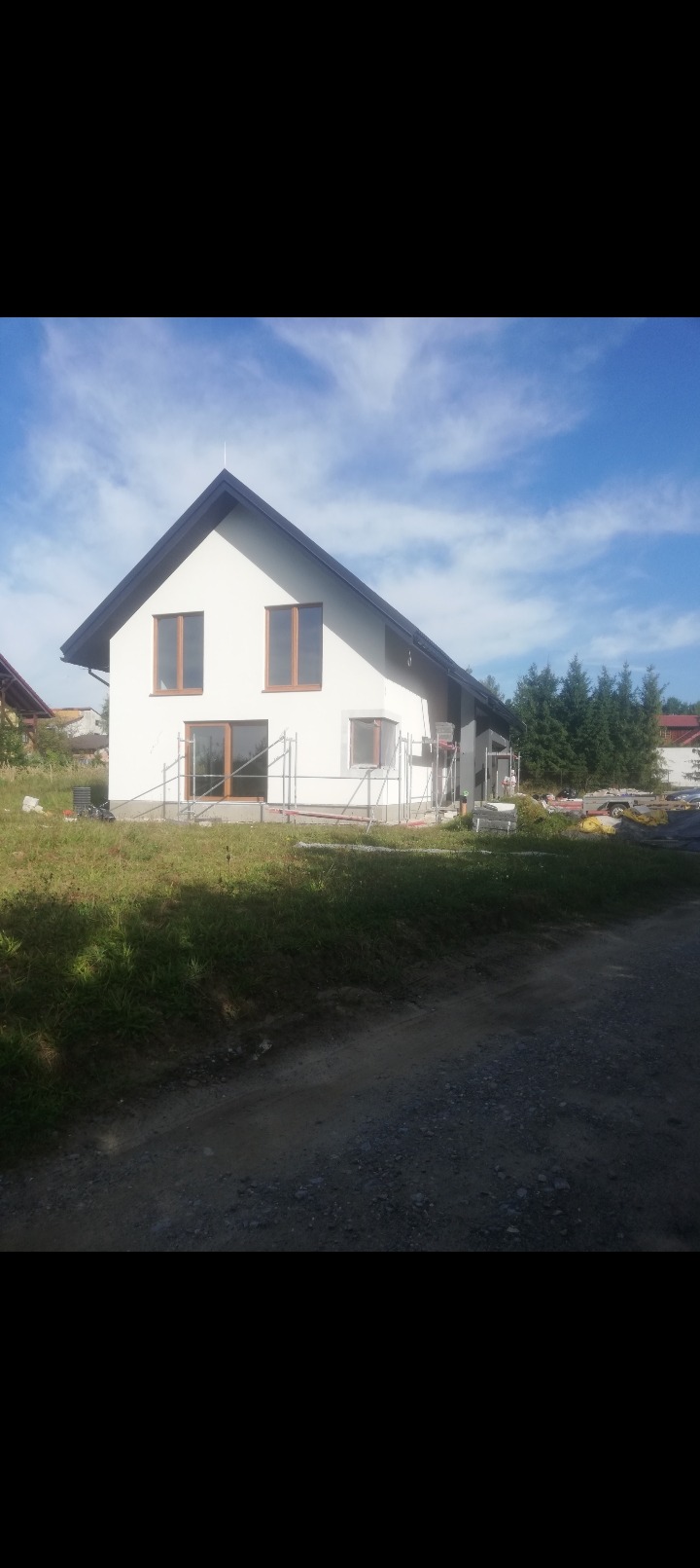 Dom jednorodzinny z białą elewacją i brązowymi oknami, częściowo otoczony rusztowaniami. Widoczne materiały budowlane i drzewa w tle. Ujęcie z dołu.