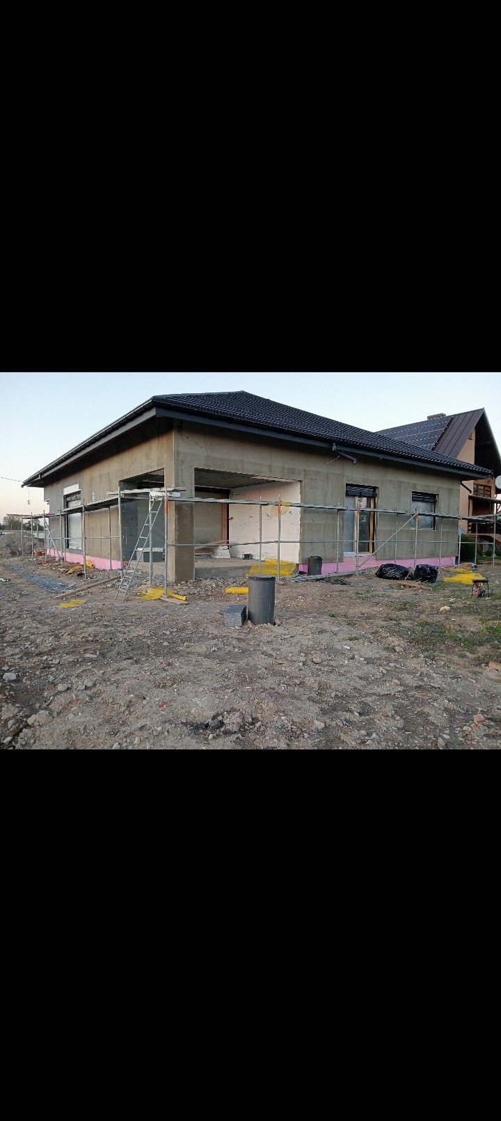 Dom w trakcie ocieplania elewacji, widoczne rusztowania i warstwa izolacyjna. Budynek z ciemnym dachem w surowym stanie, przygotowany do dalszych prac wykończeniowych.