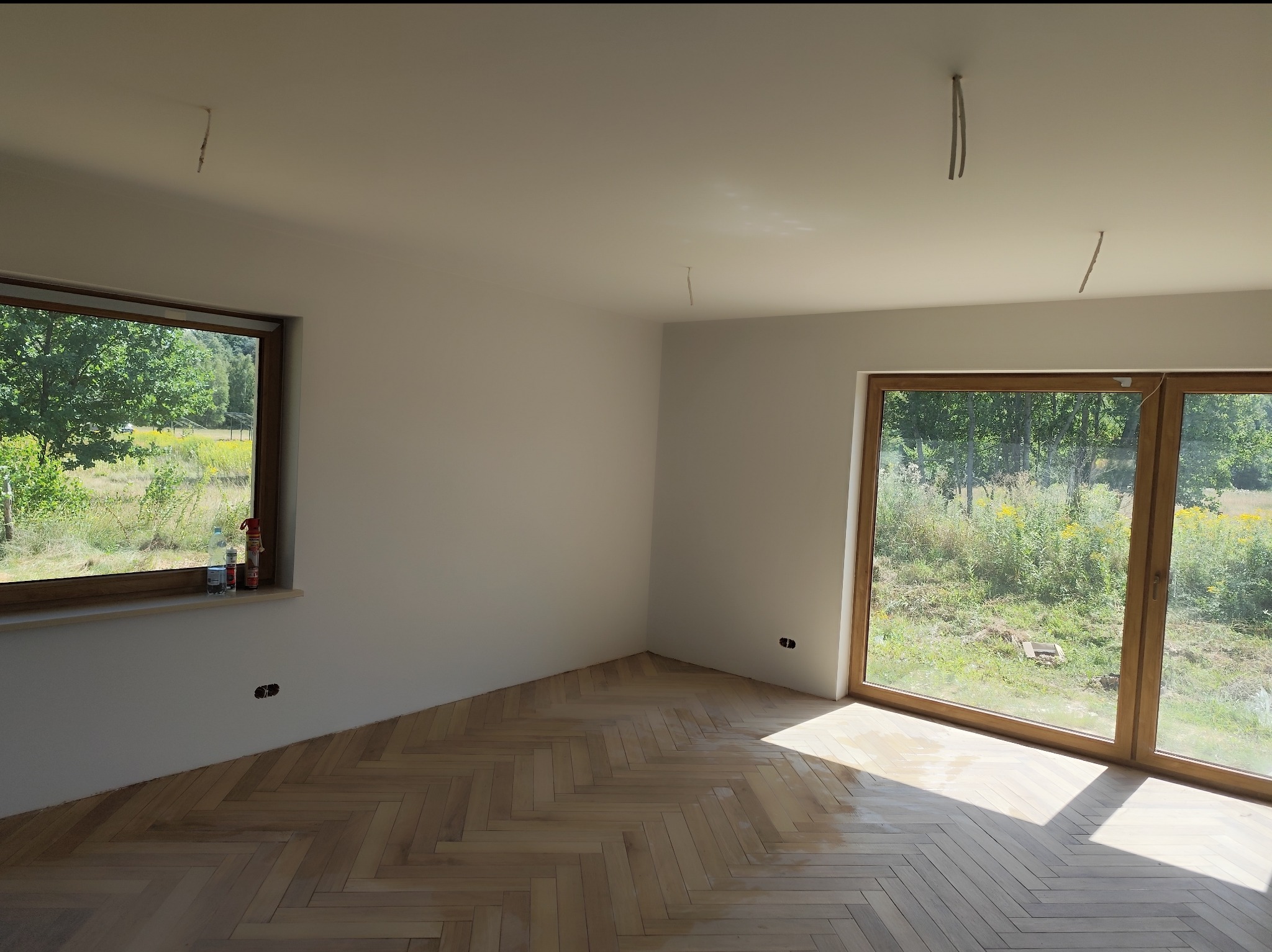 Jasne, wykończone wnętrze z parkietem ułożonym w jodełkę. Widoczne okna z drewnianą ramą i odsłonięte przewody na suficie. Świeżo wykończone ściany.