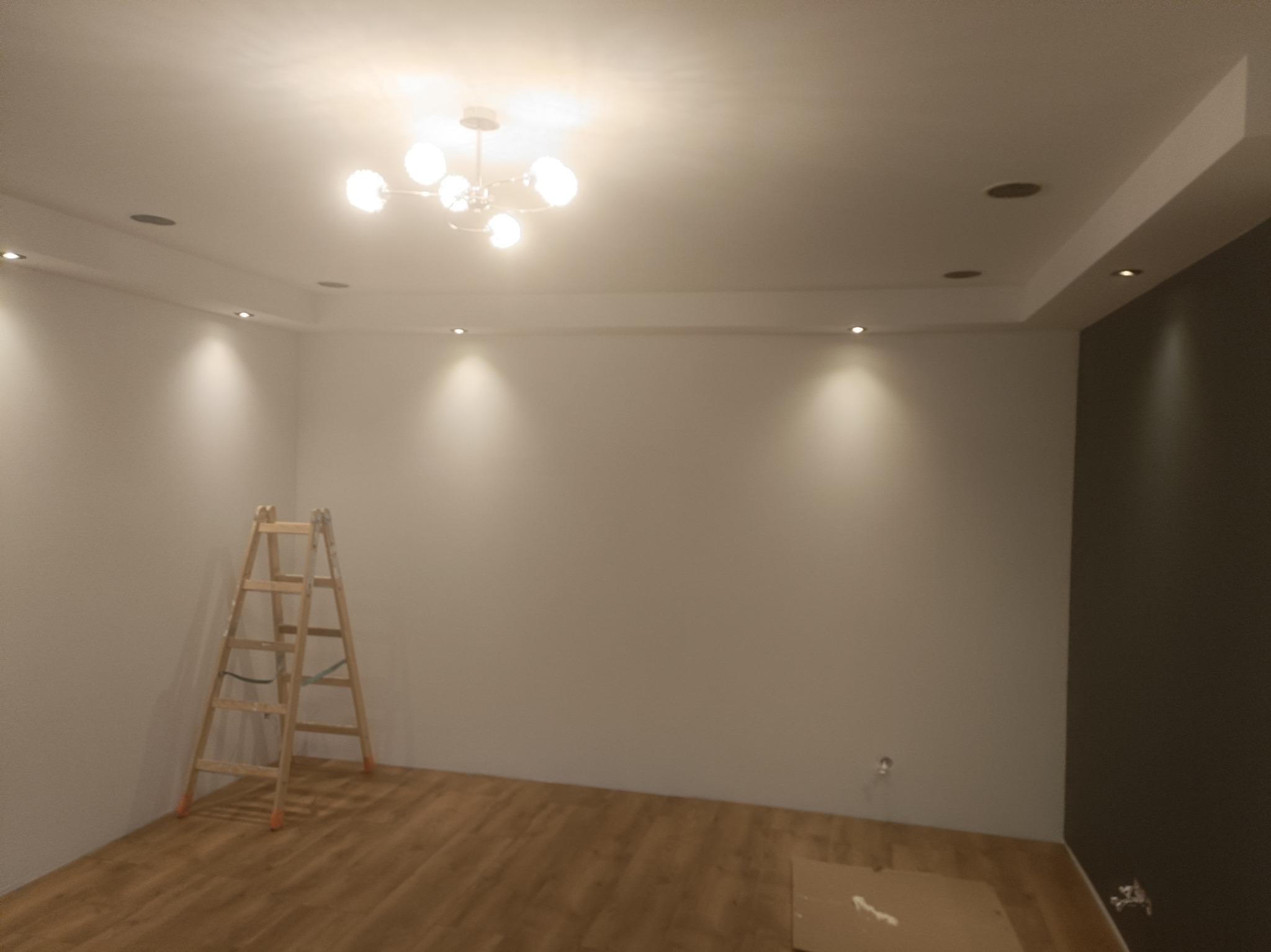 Pokój po malowaniu z drewnianą podłogą, widoczna drabina i oświetlenie sufitowe. Ściany w odcieniach bieli i szarości, z zamontowanymi punktowymi lampami.