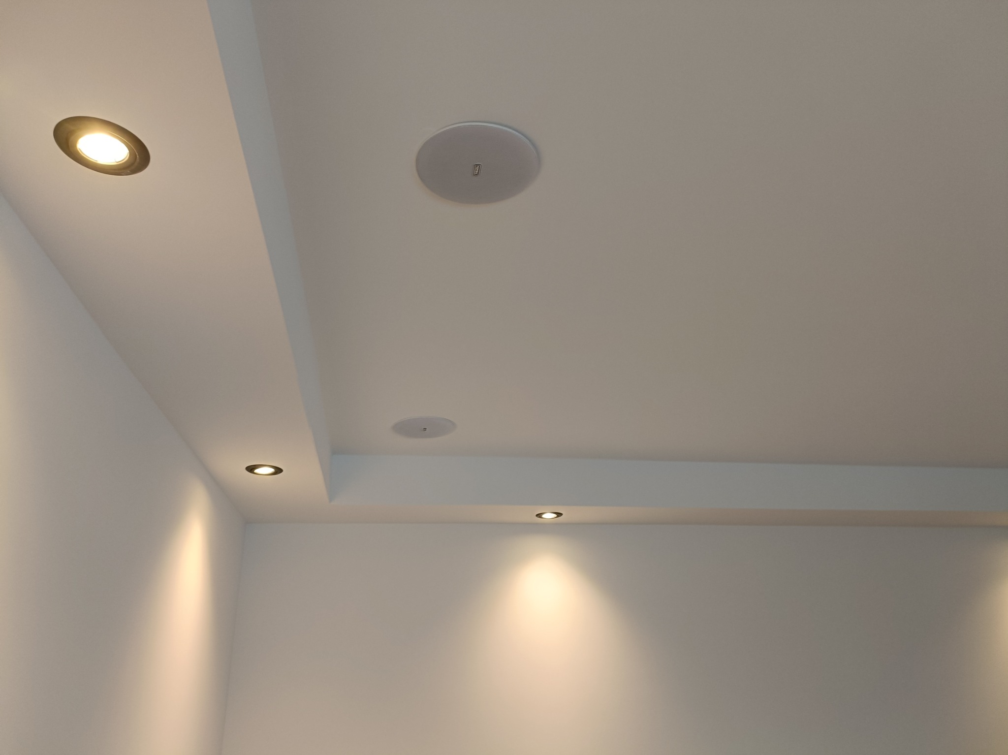 Biały sufit podwieszany z zamontowanymi okrągłymi lampami halogenowymi i głośnikiem sufitowym, oświetlający gładkie ściany w jasnym wnętrzu.