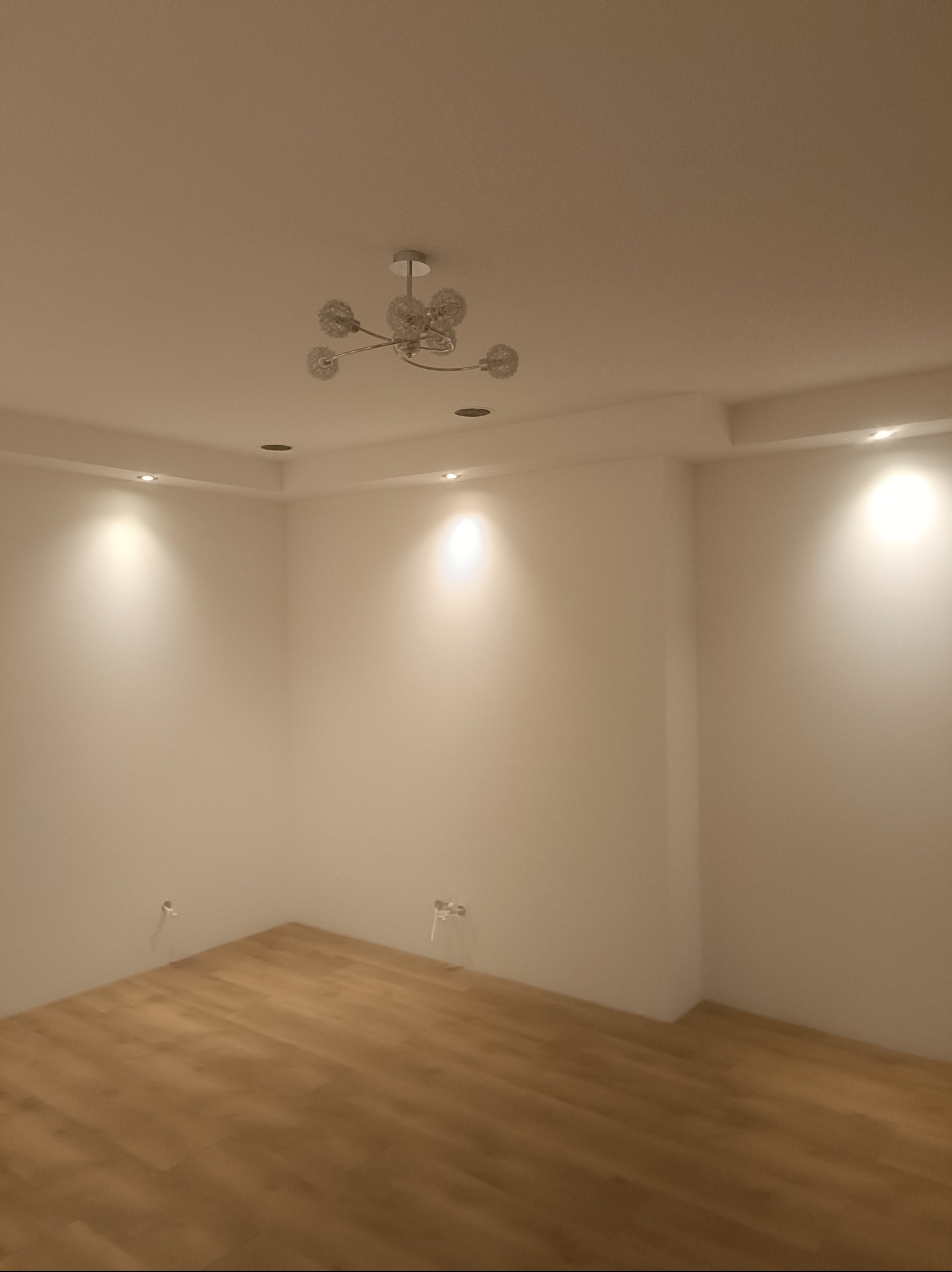 Jasne, puste wnętrze z drewnianą podłogą i nowoczesnym oświetleniem sufitowym. Widoczne punkty świetlne w suficie i wystające kable elektryczne.