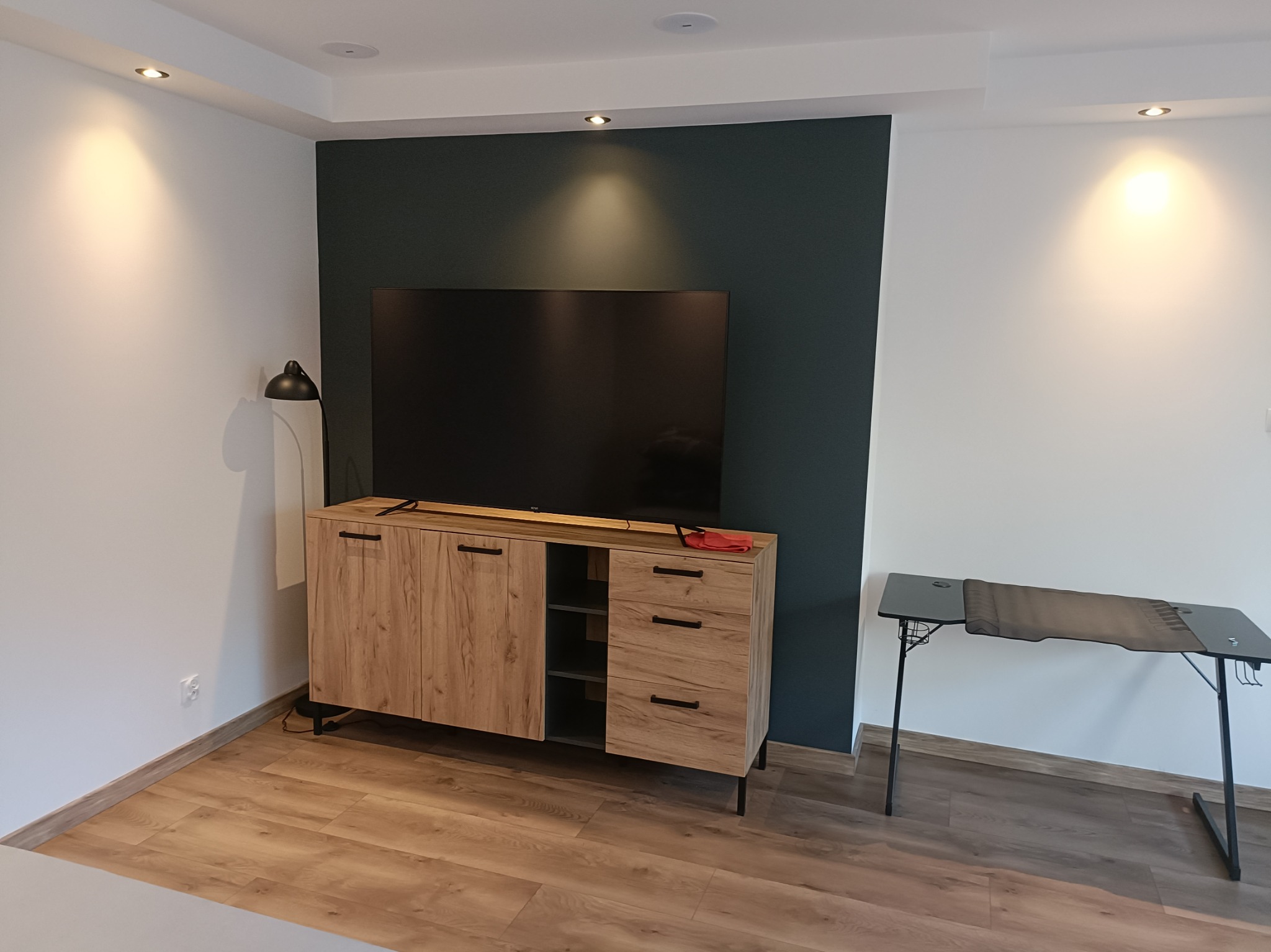 Nowoczesne wykończenie wnętrza salonu z drewnianą komodą pod telewizor i panelami podłogowymi w odcieniach brązu, oświetlenie punktowe.