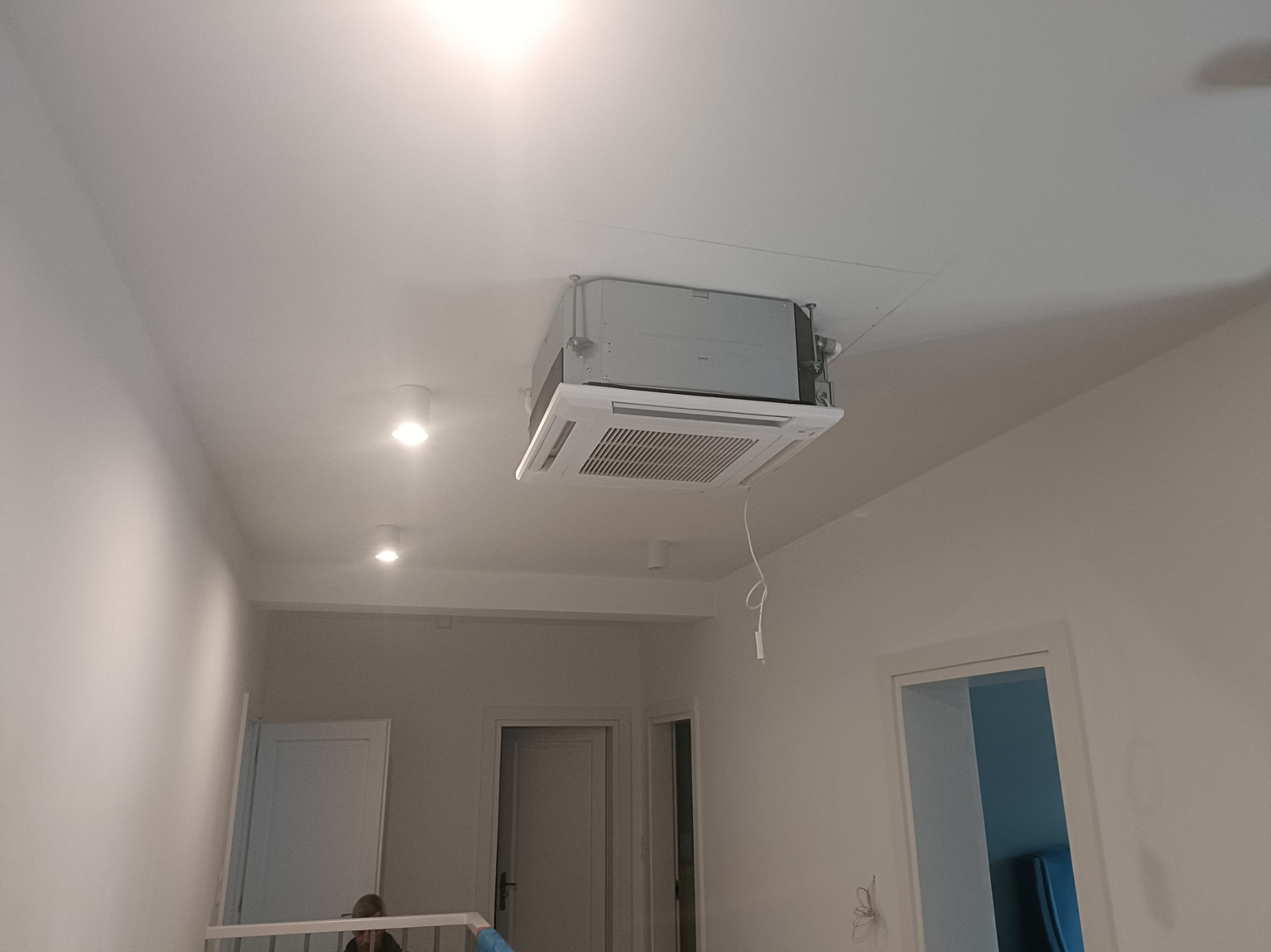 Kasetonowe klimatyzacja sufitowa w jasnym wnętrzu. Widoczne elementy montażowe i przewody. Białe ściany, drzwi w tle, oświetlenie punktowe.