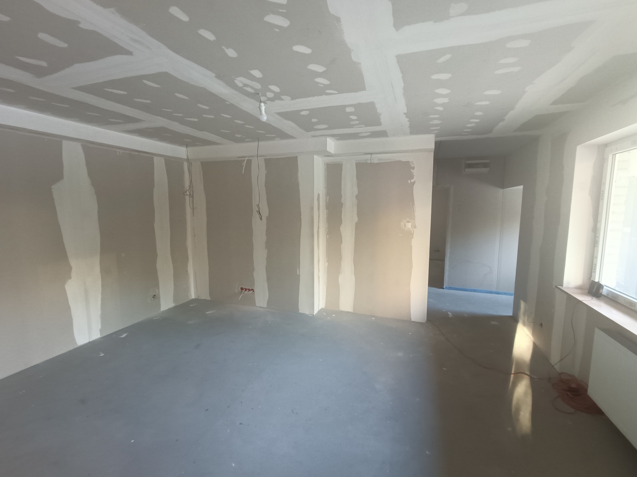Surowe wnętrze po szpachlowaniu: ściany i sufit pokryte gładzią, widoczne łączenia płyt. Minimalistyczny wystrój, przygotowanie do dalszych prac wykończeniowych.