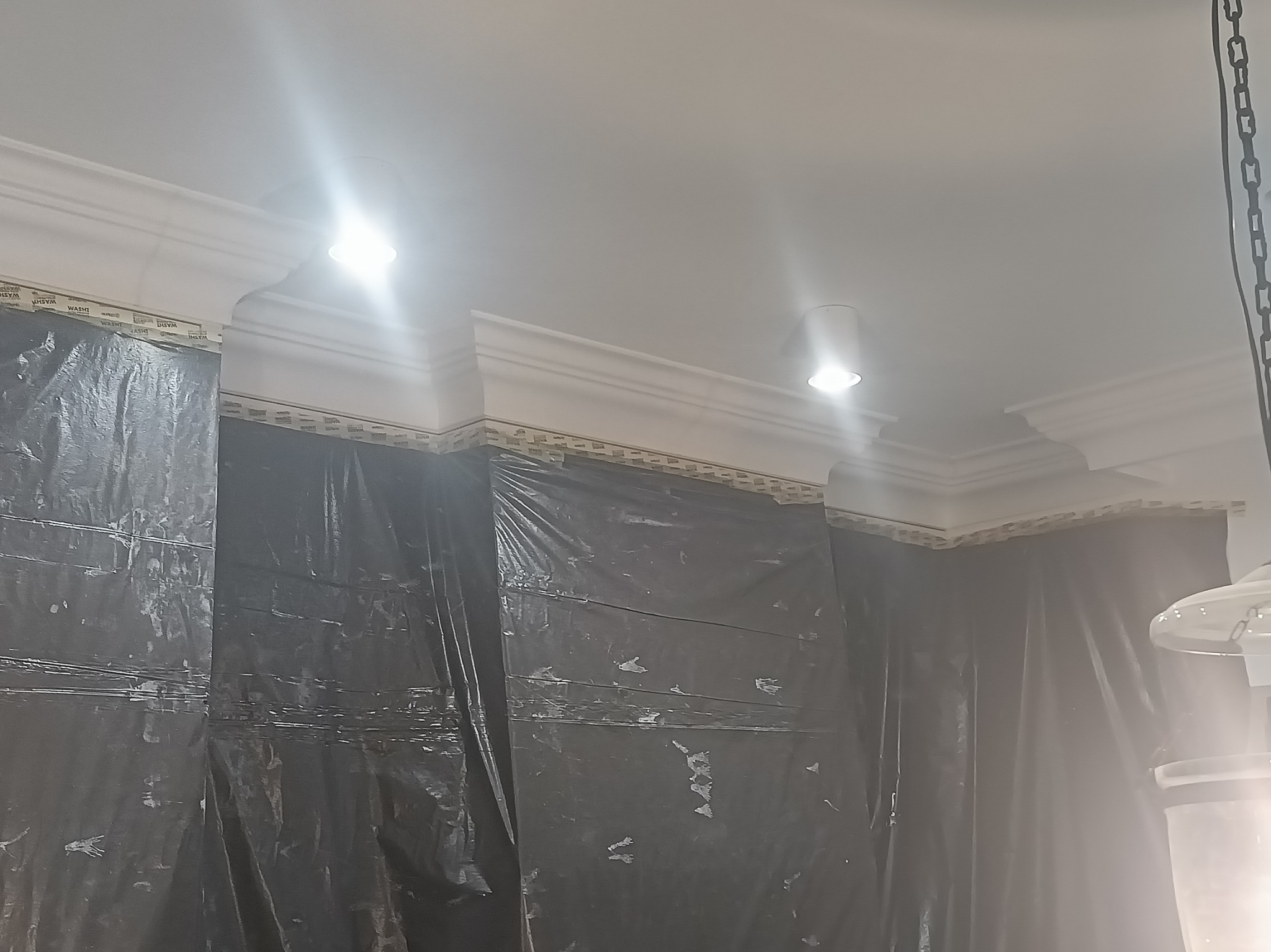 Przygotowanie do malowania: ściany osłonięte folią malarską, widoczne listwy dekoracyjne pod sufitem i oświetlenie punktowe. Etap prac wykończeniowych.