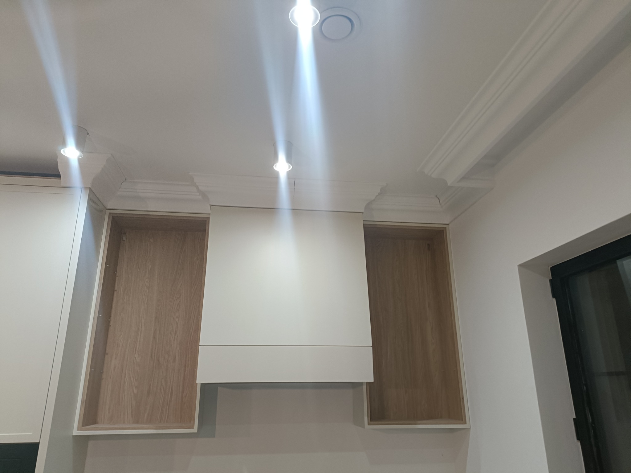 Biała zabudowa kuchenna z dekoracyjnymi listwami sufitowymi i oświetleniem punktowym. Widoczne drewniane wnętrza szafek i fragment okna.