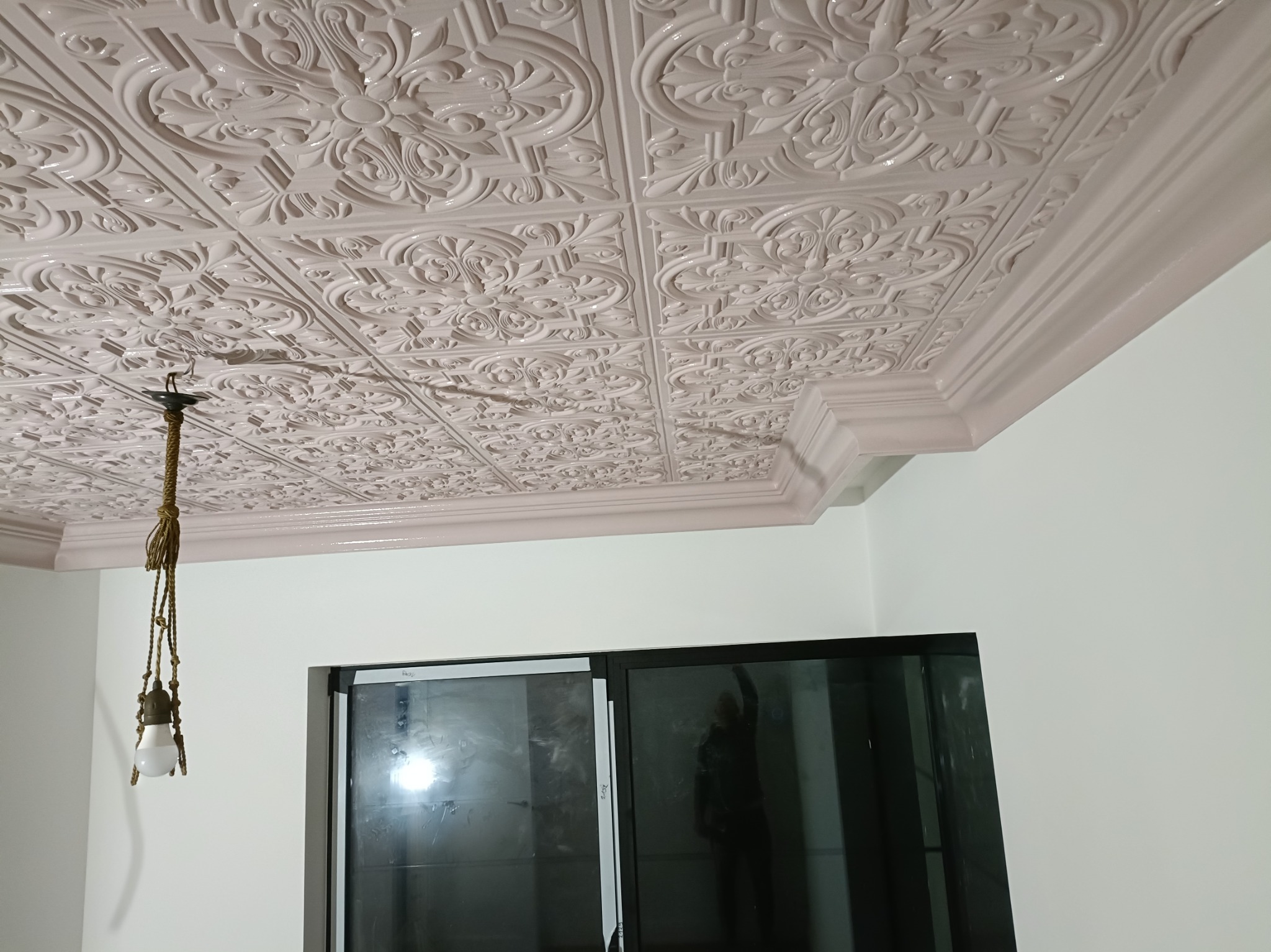 Dekoracyjny sufit z kasetonów w odcieniach bieli i beżu, wykończony listwą przysufitową. Widoczna lampa wisząca i fragment okna.