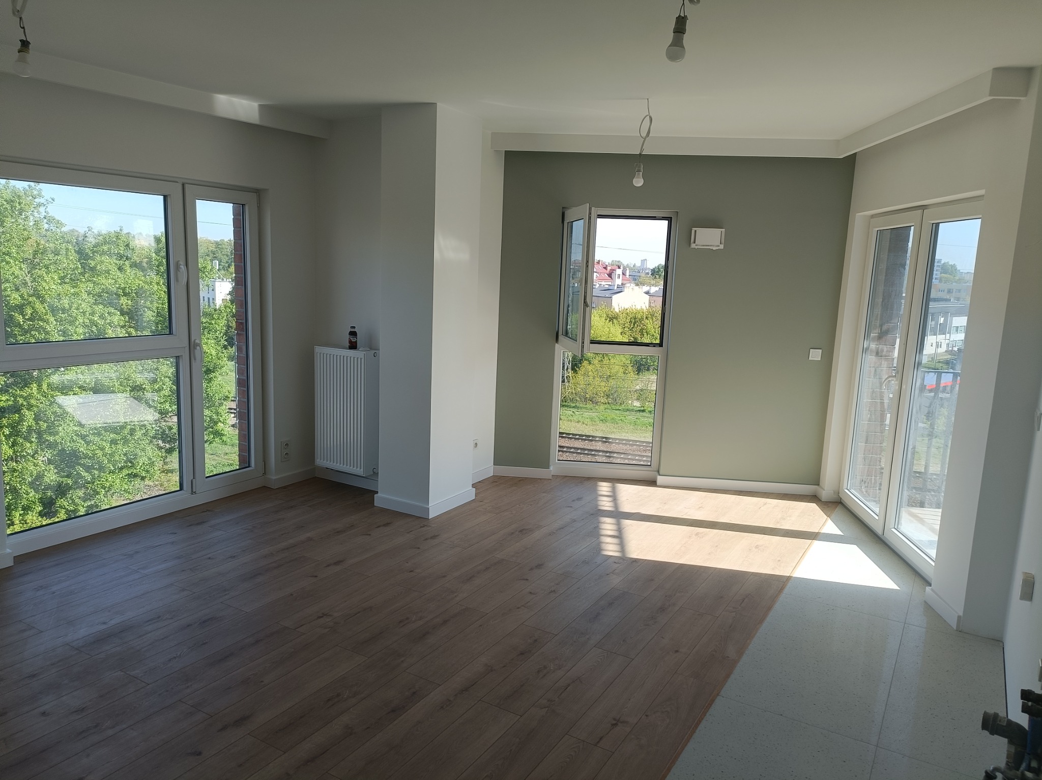Jasne, przestronne wnętrze po remoncie: drewniane panele podłogowe, białe ściany i okna z widokiem na zieleń. Część podłogi jeszcze nieukończona.