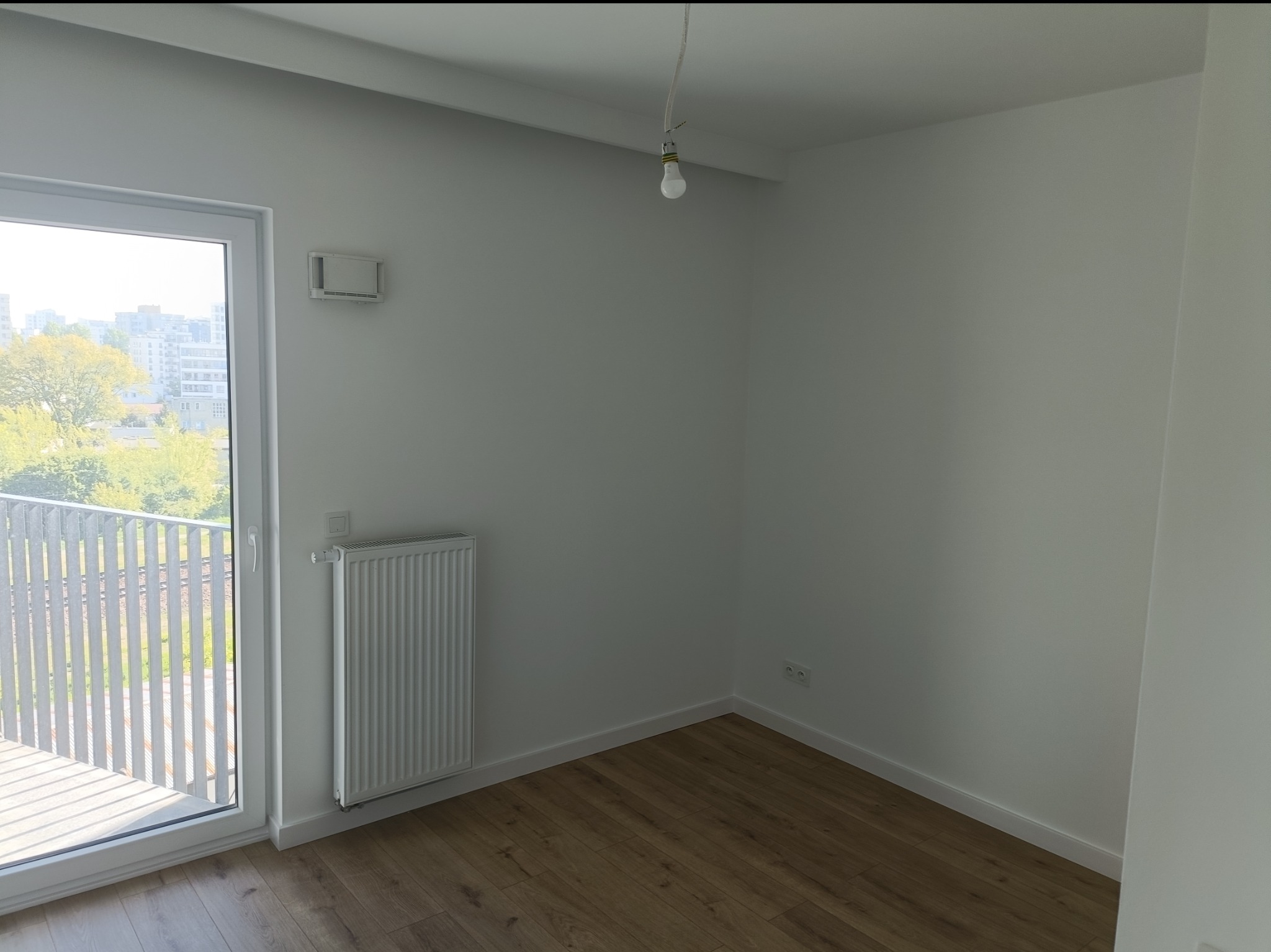 Jasne wnętrze z panelami podłogowymi i białymi ścianami. Widok na balkon z metalową balustradą i zielenią w tle. Grzejnik pod oknem, surowa żarówka.