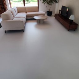 MicroArt - Jasnoszara podłoga w salonie z narożną sofą, okrągłym stolikiem kawowym z marmurowym blatem i drewnianą szafką RTV, duża roślina doniczkowa obok okna.