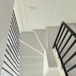 MicroArt - Minimalistyczne, białe schody z mikrocementu z czarną balustradą i marmurową podłogą, widziane z góry, oświetlone naturalnym światłem.