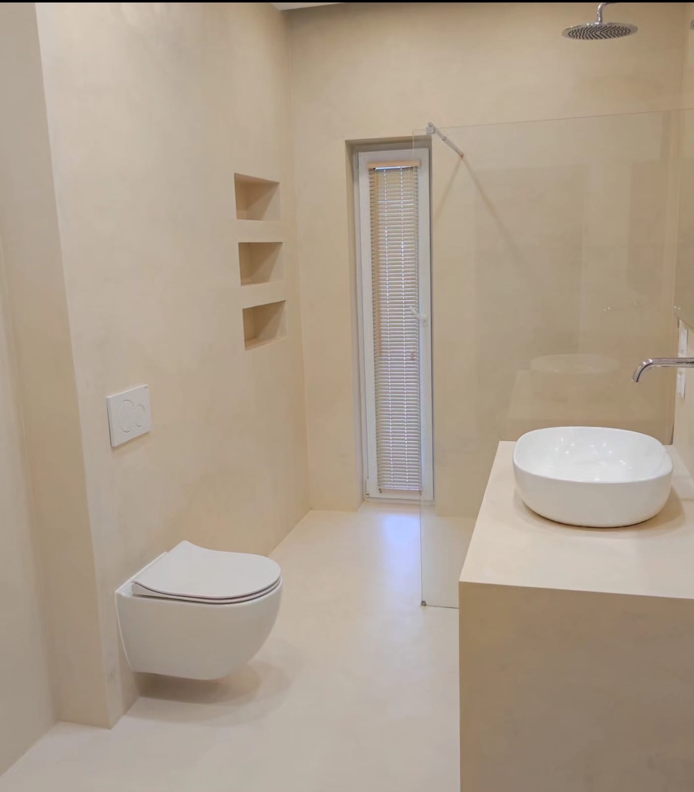 Beżowa łazienka z mikrocementem na ścianach i podłodze, minimalistyczna umywalka nablatowa, podwieszana toaleta i kabina prysznicowa ze szklaną ścianką.