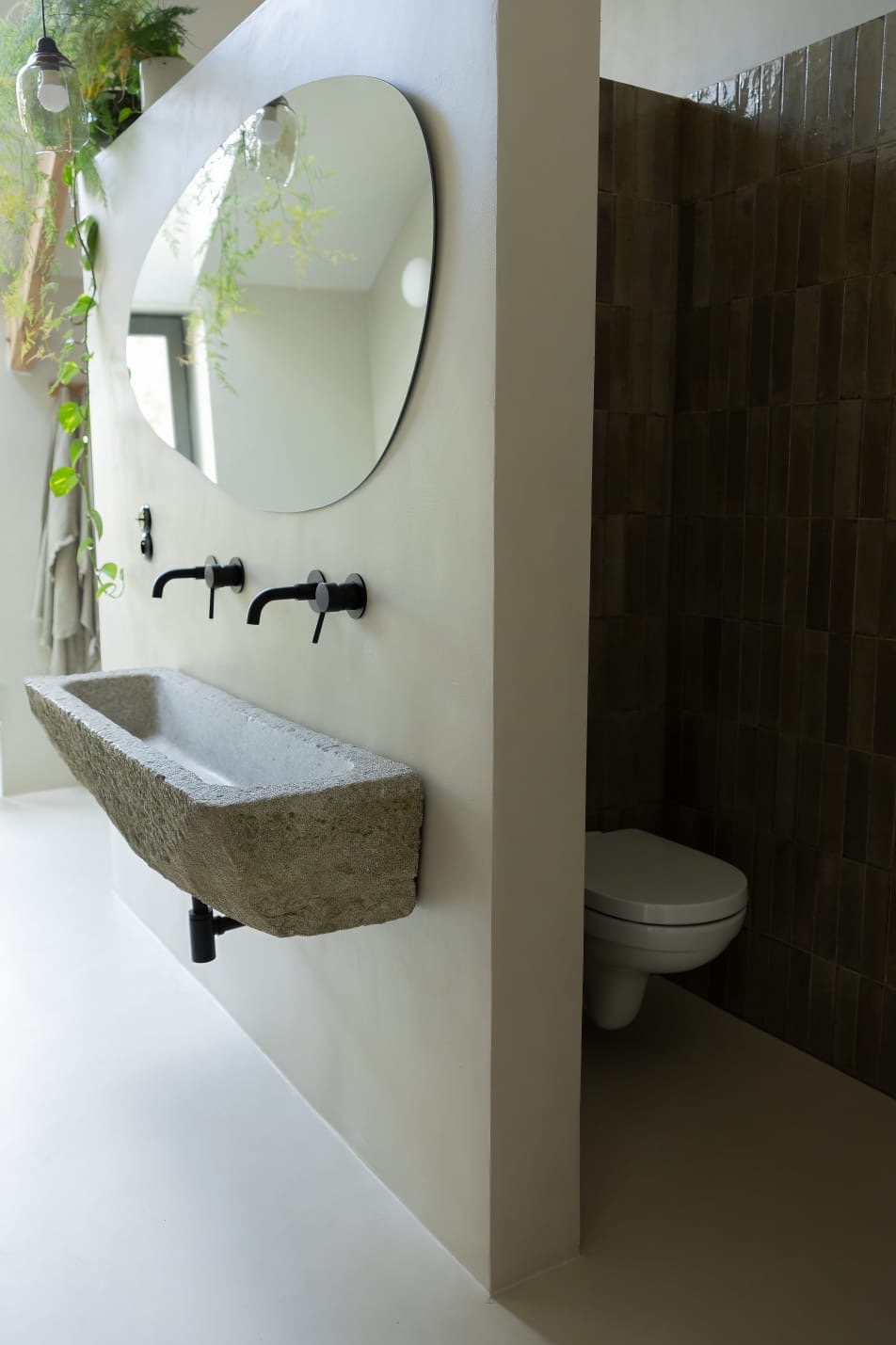 Minimalistyczna łazienka z kamienną umywalką, czarną armaturą i owalnym lustrem na ścianie wykończonej w jasnym odcieniu, widoczny fragment toalety z ciemnymi płytkami w tle.