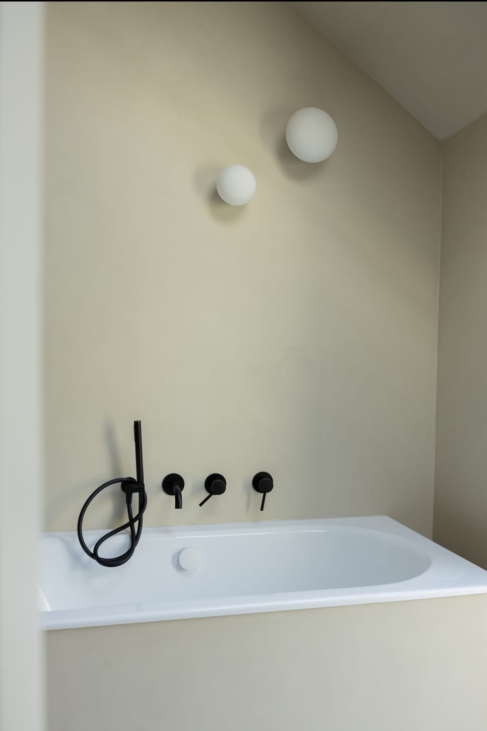 Minimalistyczna łazienka z białą wanną, czarną armaturą i dwoma okrągłymi lampami na beżowej ścianie.
