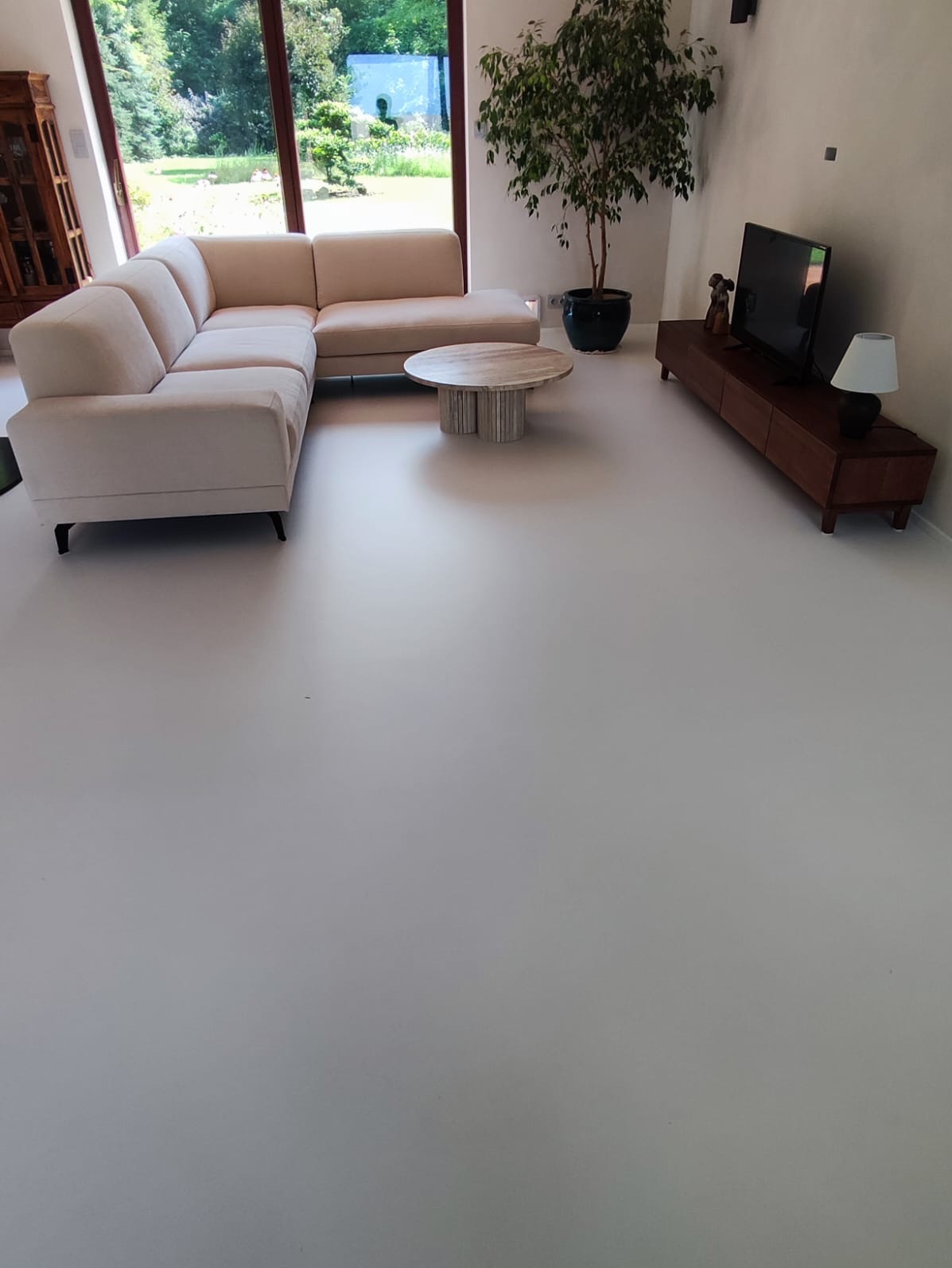 Jasnoszara podłoga w salonie z narożną sofą, okrągłym stolikiem kawowym z marmurowym blatem i drewnianą szafką RTV, duża roślina doniczkowa obok okna.