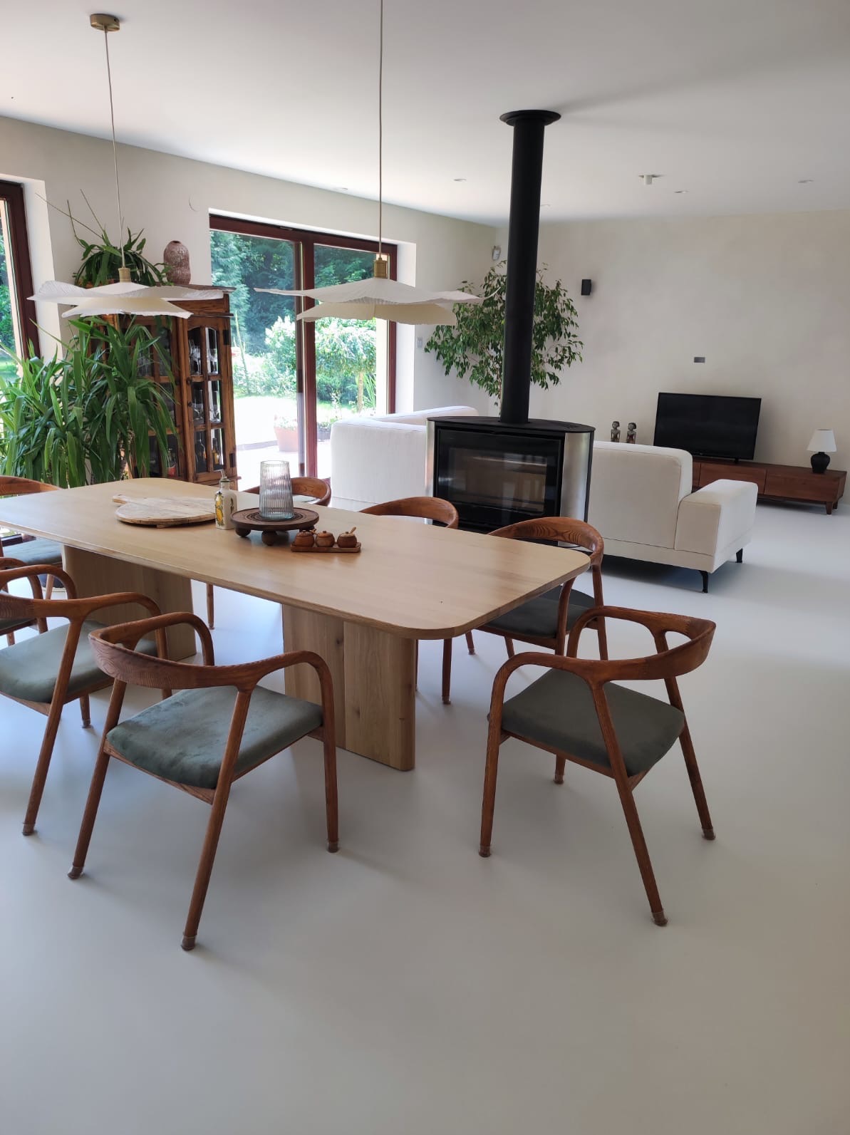 Jasne wnętrze z drewnianym stołem i krzesłami z zielonymi siedziskami, kominek z czarnym kominem, rośliny doniczkowe i biała podłoga w minimalistycznym stylu.