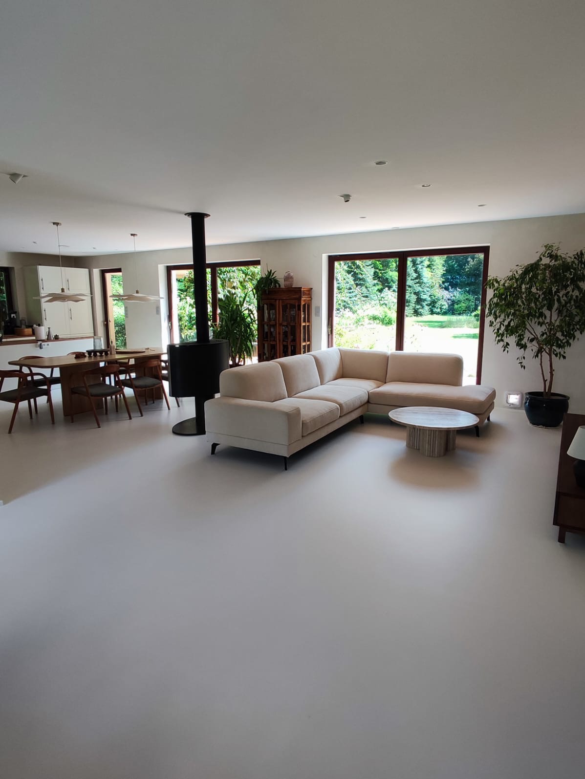 Jasnoszara podłoga z mikrocementu w przestronnym salonie z narożną sofą, okrągłym stolikiem kawowym i czarnym piecem wolnostojącym.