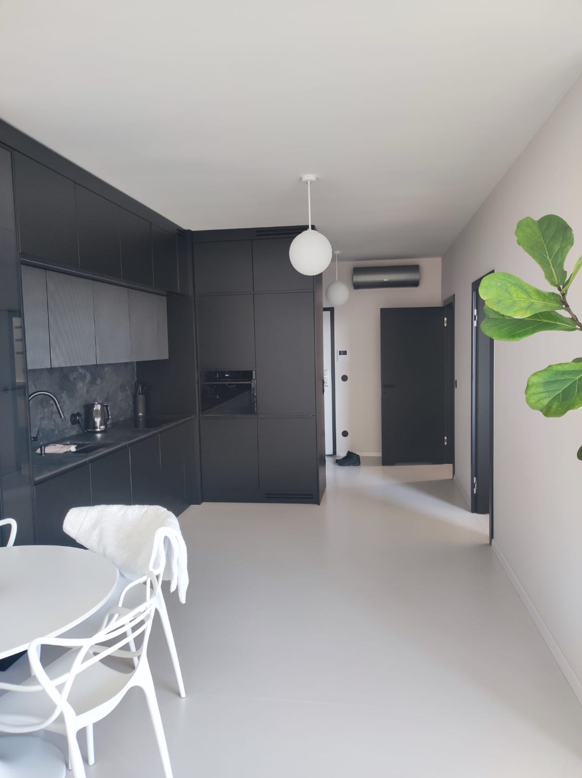 Nowoczesna kuchnia z czarnymi szafkami i jasnoszarą podłogą, oświetlona kulistymi lampami, z widocznym fragmentem stołu i krzeseł oraz rośliną doniczkową po prawej stronie.