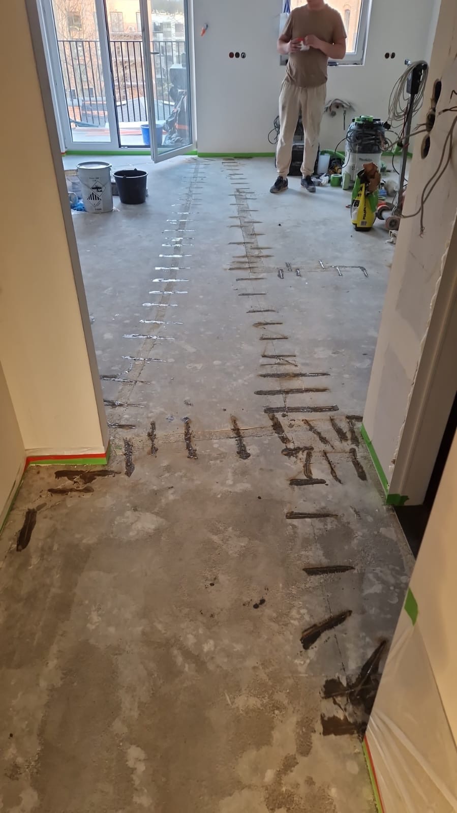 Przygotowanie podłoża pod posadzkę: widoczne nacięcia w betonie i zabezpieczone taśmą malarską ściany, w tle balkon i osoba w jasnych spodniach.