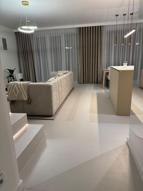 Jasna, minimalistyczna posadzka w salonie z beżową sofą i schodami z podświetleniem LED. Nowoczesne lampy wiszące i zasłony w tle.