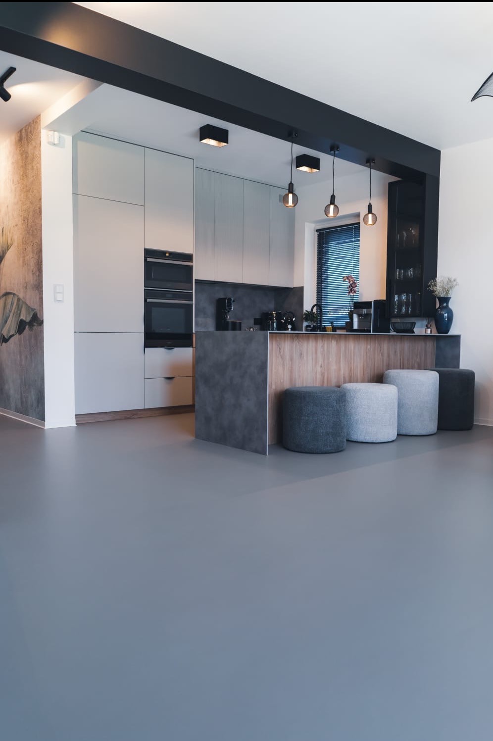 Szara, gładka posadzka w nowoczesnej kuchni z wyspą i hokerami. Minimalistyczne wnętrze z akcentami drewna i ciemnymi belkami sufitowymi.