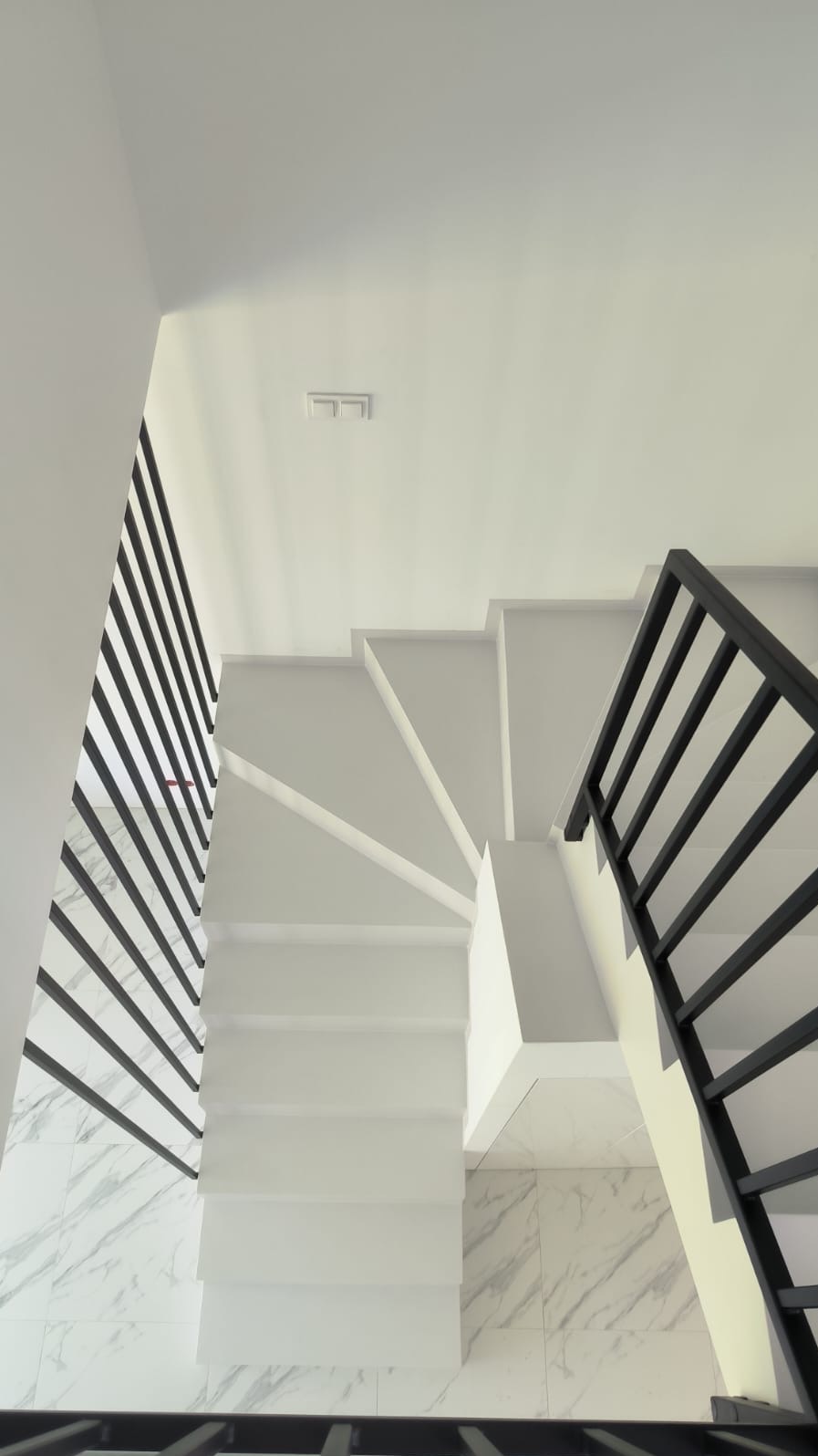 Minimalistyczne, białe schody z mikrocementu z czarną balustradą i marmurową podłogą, widziane z góry, oświetlone naturalnym światłem.