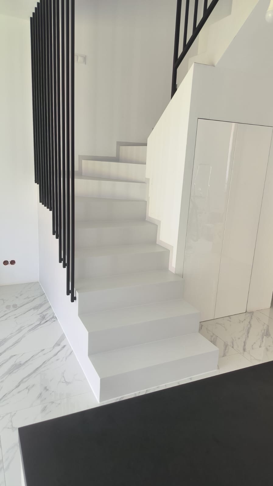 Minimalistyczne, białe schody z czarną balustradą w nowoczesnym wnętrzu. Marmurowa podłoga i białe ściany podkreślają czystość formy. Widoczne gniazdka elektryczne.