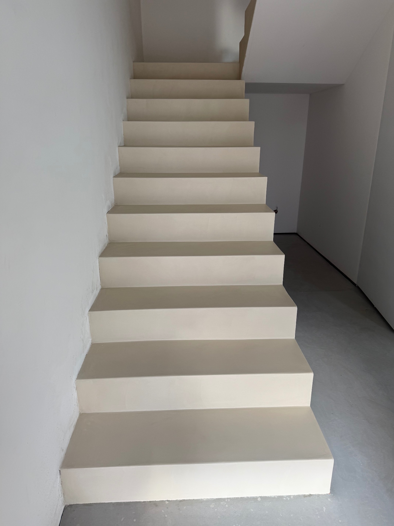 Schody wewnętrzne wykończone jednolitą, jasną powłoką, prawdopodobnie żywiczną. Minimalistyczny design, gładkie powierzchnie, widok z dołu. Nowoczesne wykończenie.