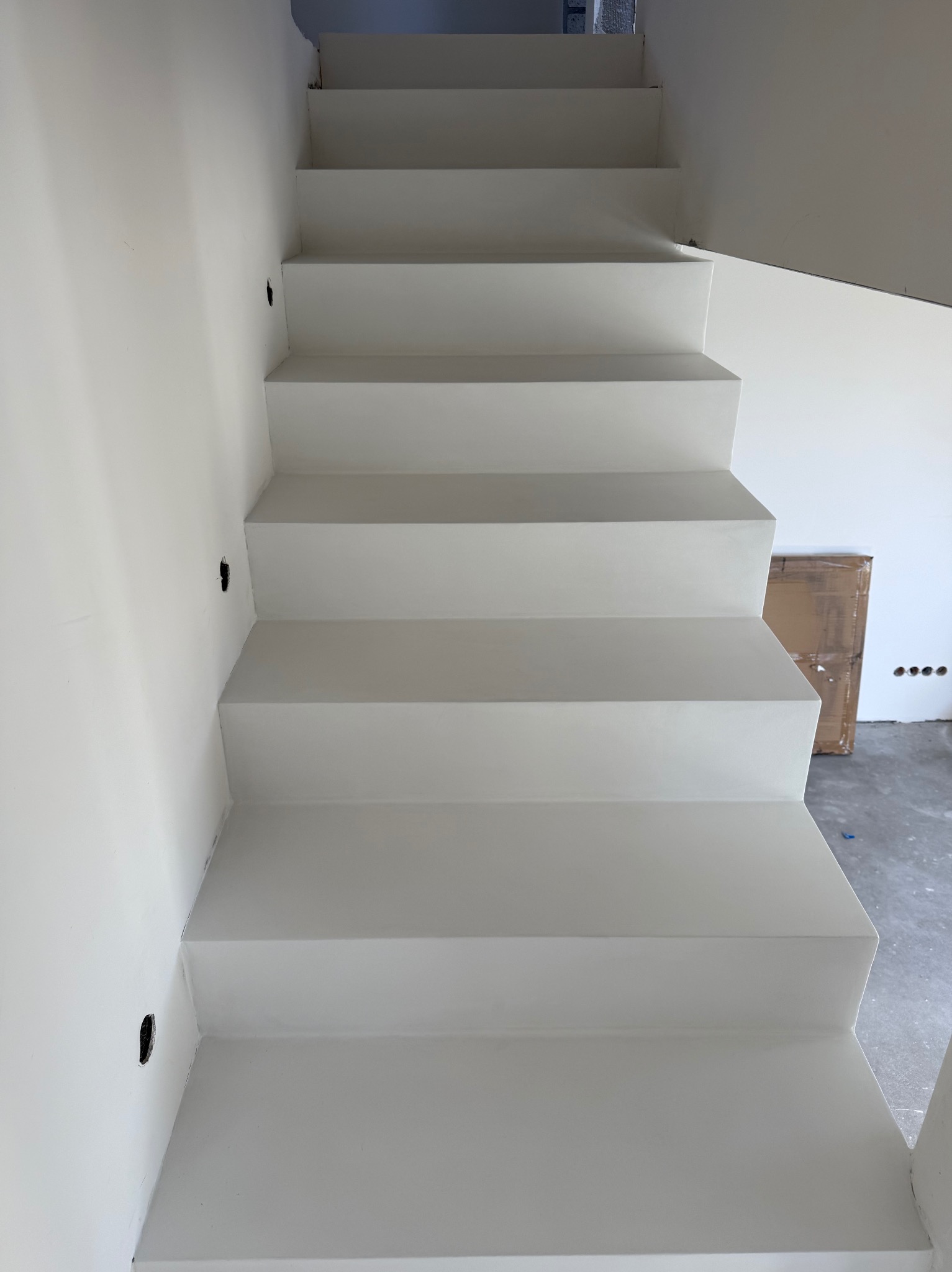 Białe schody z równymi stopniami, widok z dołu, minimalistyczny design, ściana z otworami na oświetlenie, karton w tle, surowy beton na podłodze.
