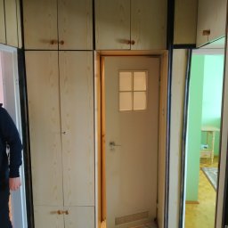 Piotr Malinowski - Wnęka z zabudową szaf i drzwiami w odcieniach beżu i brązu, widok na sąsiednie pomieszczenie z zieloną ścianą i jasnymi meblami.