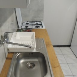 Piotr Malinowski - Kątowe ujęcie zlewu ze stali nierdzewnej z baterią i ociekaczem na drewnianym blacie, obok kuchenka gazowa w jasnej kuchni z płytkami podłogowymi.