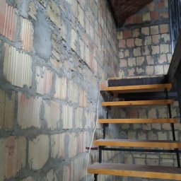 Piotr Malinowski - Surowa ściana z bloczków z widocznymi spoinami i fragmentem ocieplonego poddasza, obok schody z drewnianymi stopniami i metalową konstrukcją.