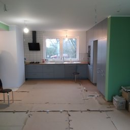 Piotr Malinowski - Wnętrze kuchni w trakcie remontu: szare szafki kuchenne, białe płytki na ścianie, podłoga zabezpieczona kartonem, ściany pomalowane na zielono, widoczne przygotowania do dalszych prac wykończeniowych.