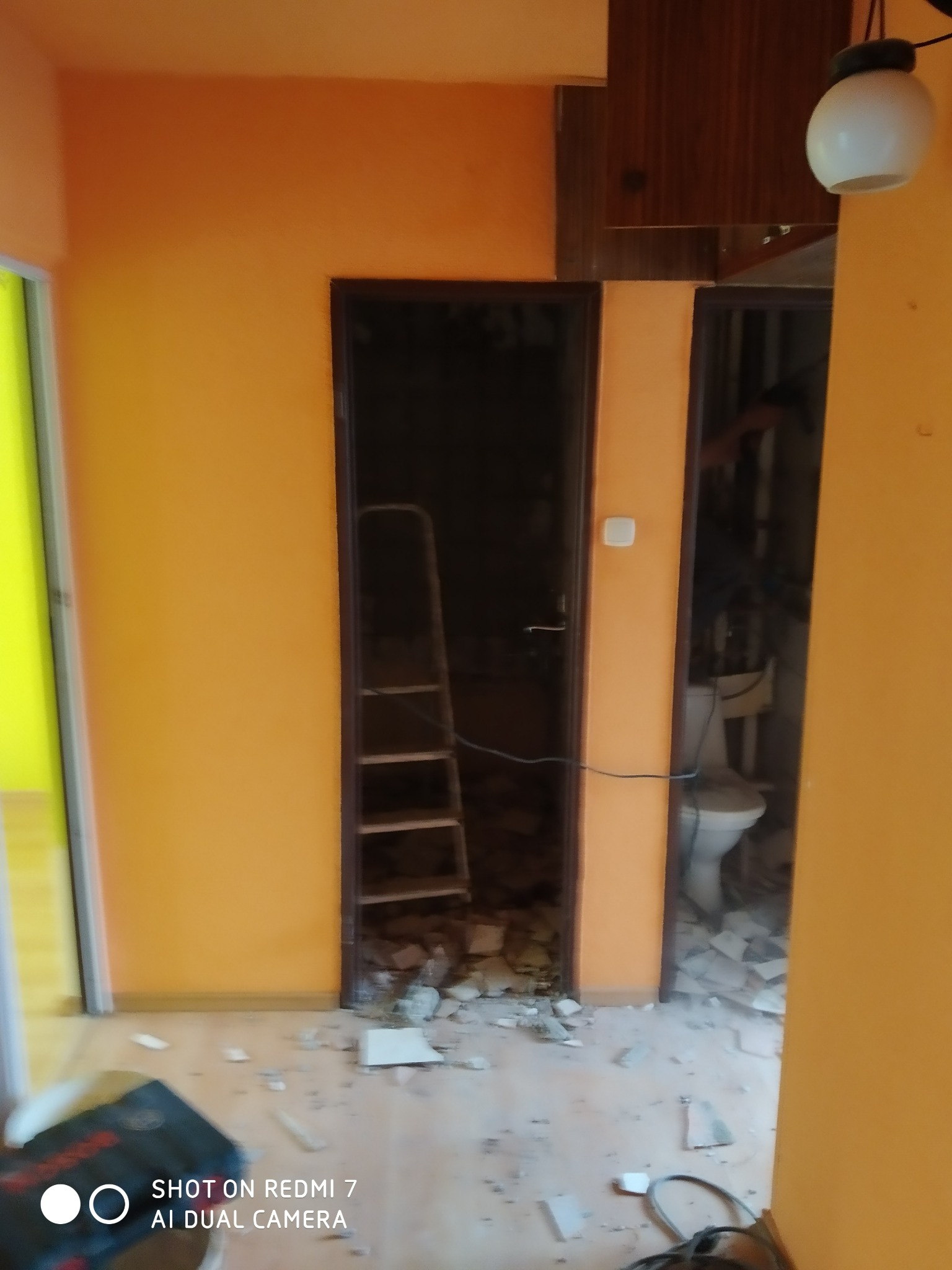 Demontaż płytek w łazience podczas remontu, widoczna drabina w otworze drzwiowym, gruz na podłodze i częściowo zdemontowana toaleta w tle.