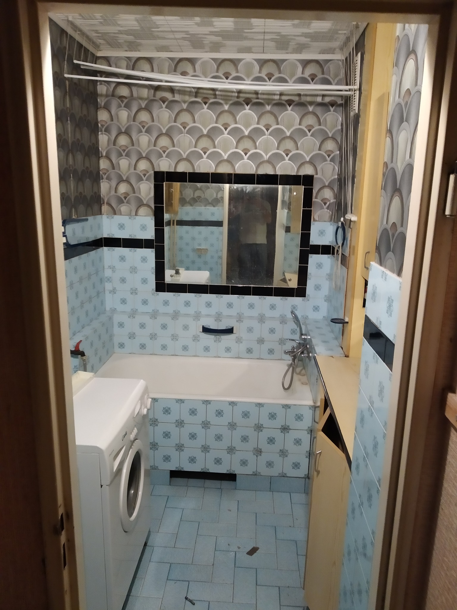 Łazienka w bloku z pralką, niebieskimi płytkami z motywem kwiatowym i tapetą w szaro-białe łuski, widok zza otwartych drzwi.