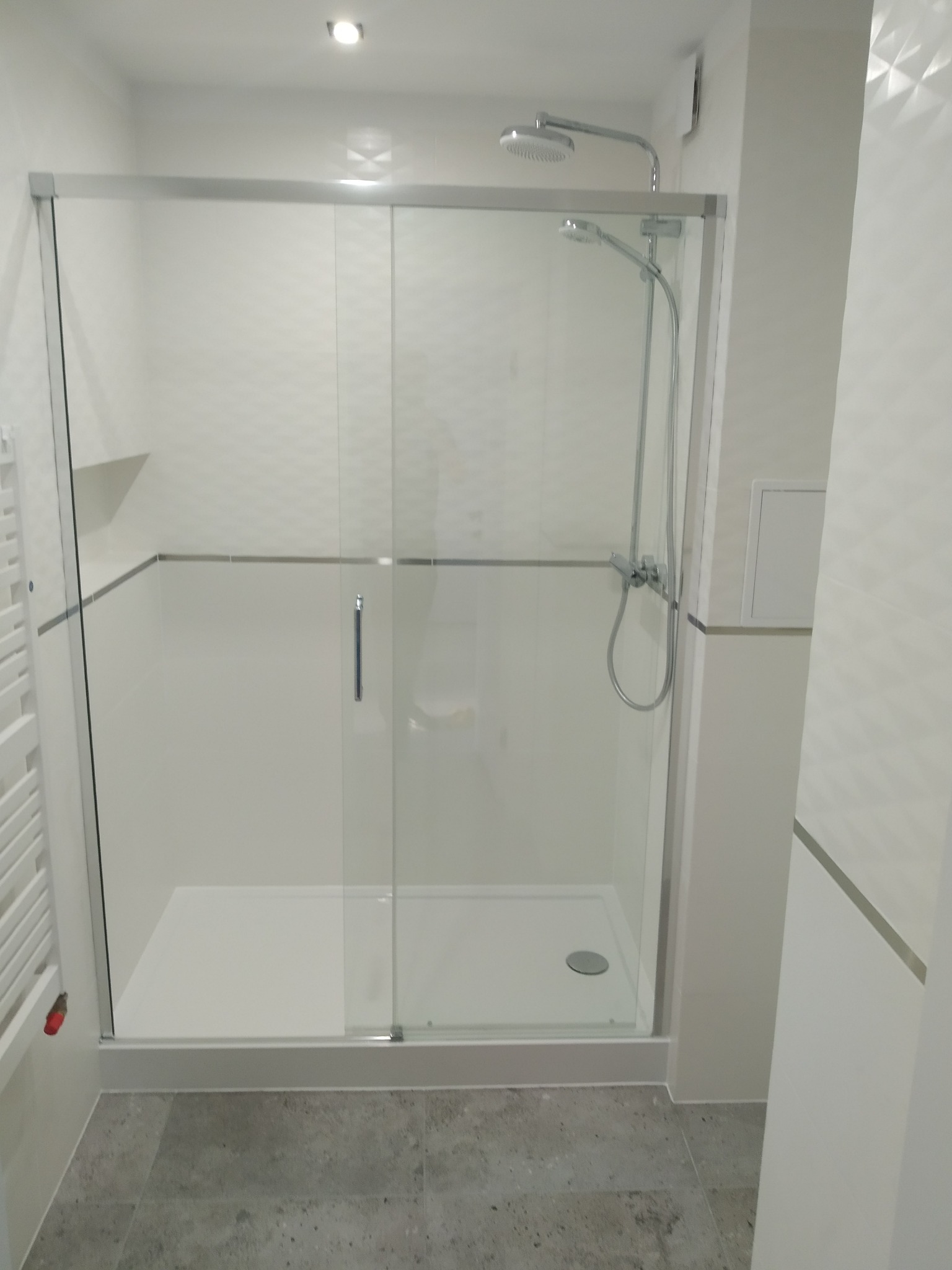 Nowoczesna kabina prysznicowa z przesuwnymi szklanymi drzwiami, zamontowana w łazience wykończonej białymi płytkami z delikatnym wzorem i szarą podłogą.