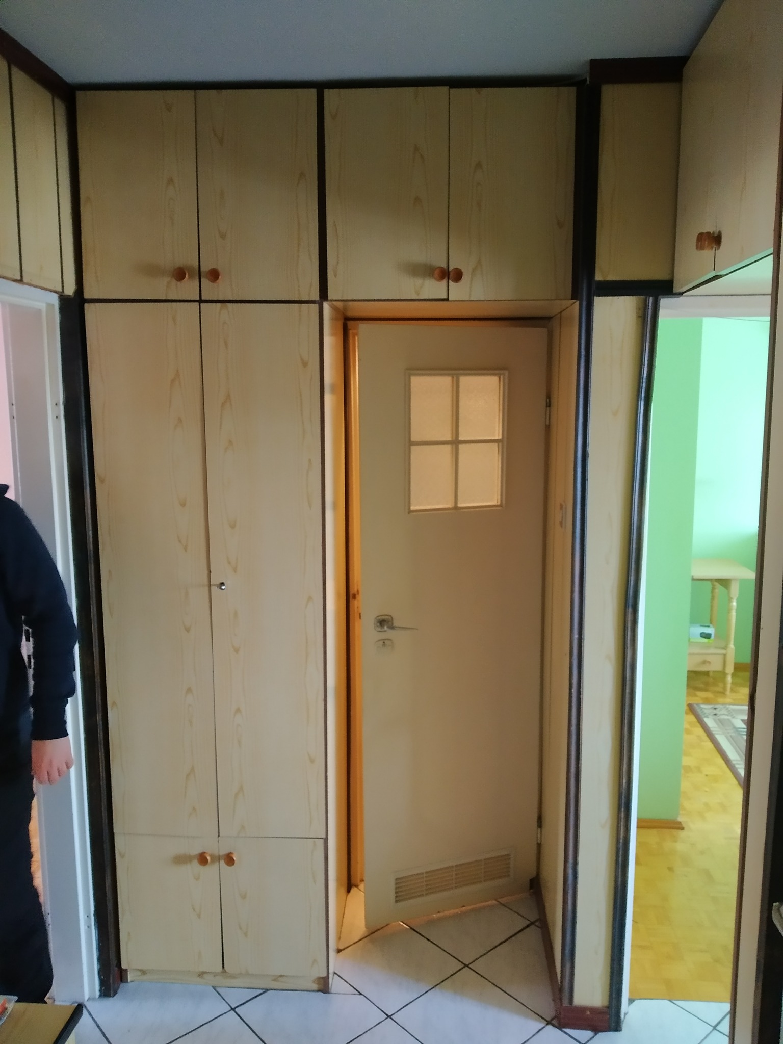 Wnęka z zabudową szaf i drzwiami w odcieniach beżu i brązu, widok na sąsiednie pomieszczenie z zieloną ścianą i jasnymi meblami.