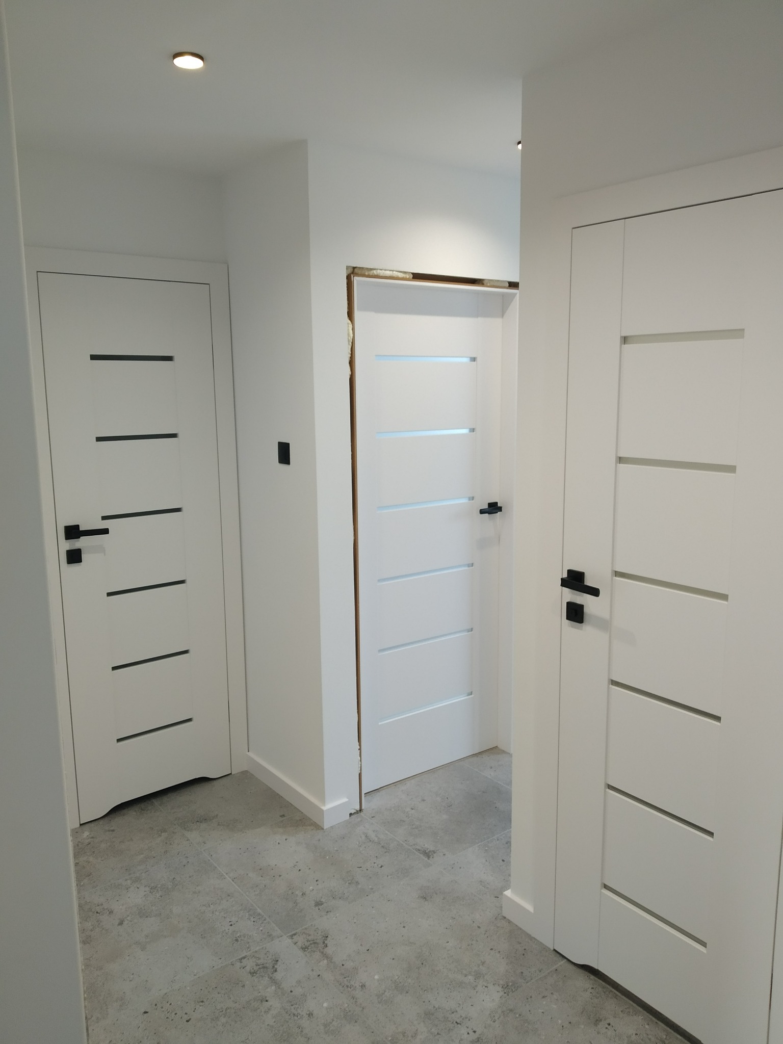 Wnętrze przedpokoju z trzema białymi drzwiami z poziomymi, czarnymi akcentami i czarnymi klamkami, szara podłoga z płytek, jedno z drzwi w trakcie montażu.