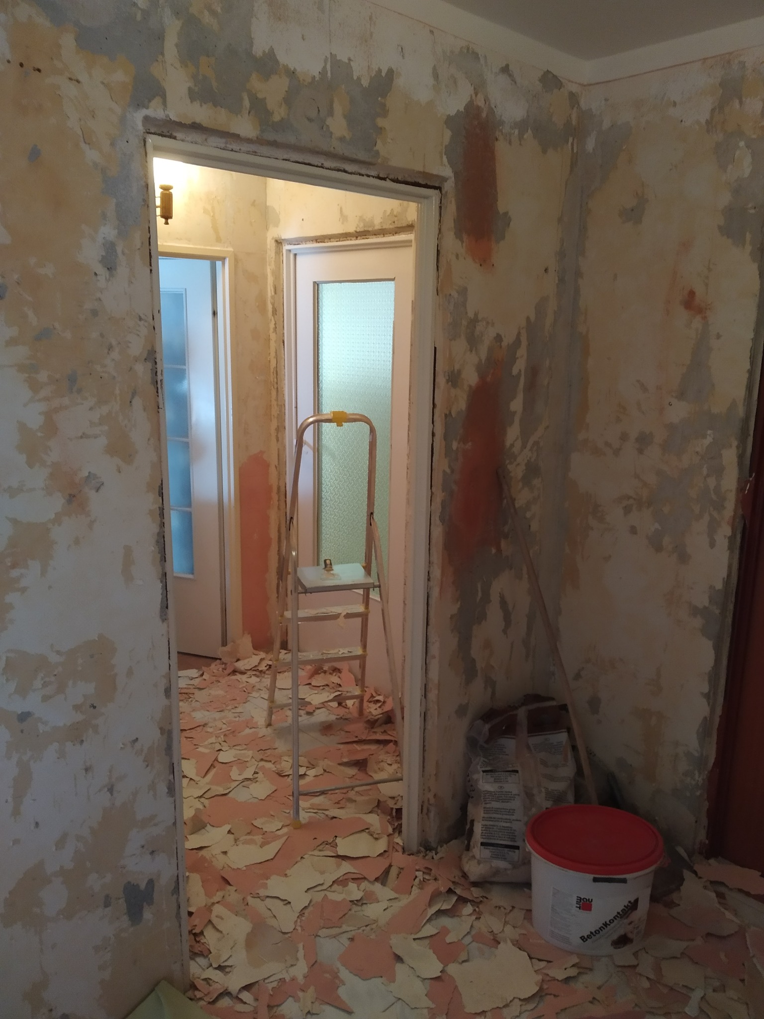 Pokój w trakcie usuwania starej tapety, z widoczną drabiną, workiem z klejem i wiadrem z napisem Betonkontakt.