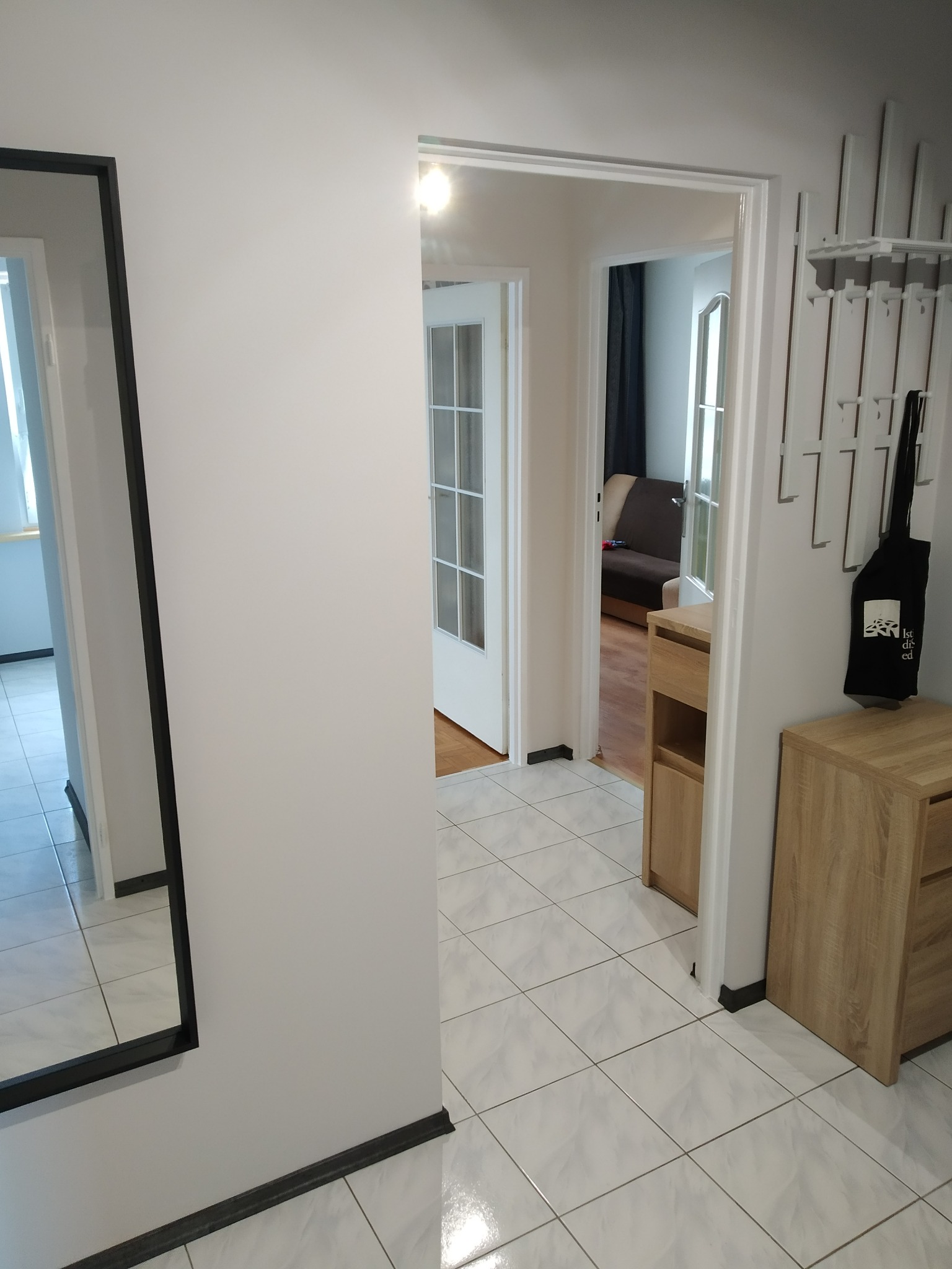 Jasne wnętrze mieszkania po remoncie: białe ściany, płytki podłogowe, minimalistyczny wieszak z torbą, widok na salon przez otwarte drzwi.