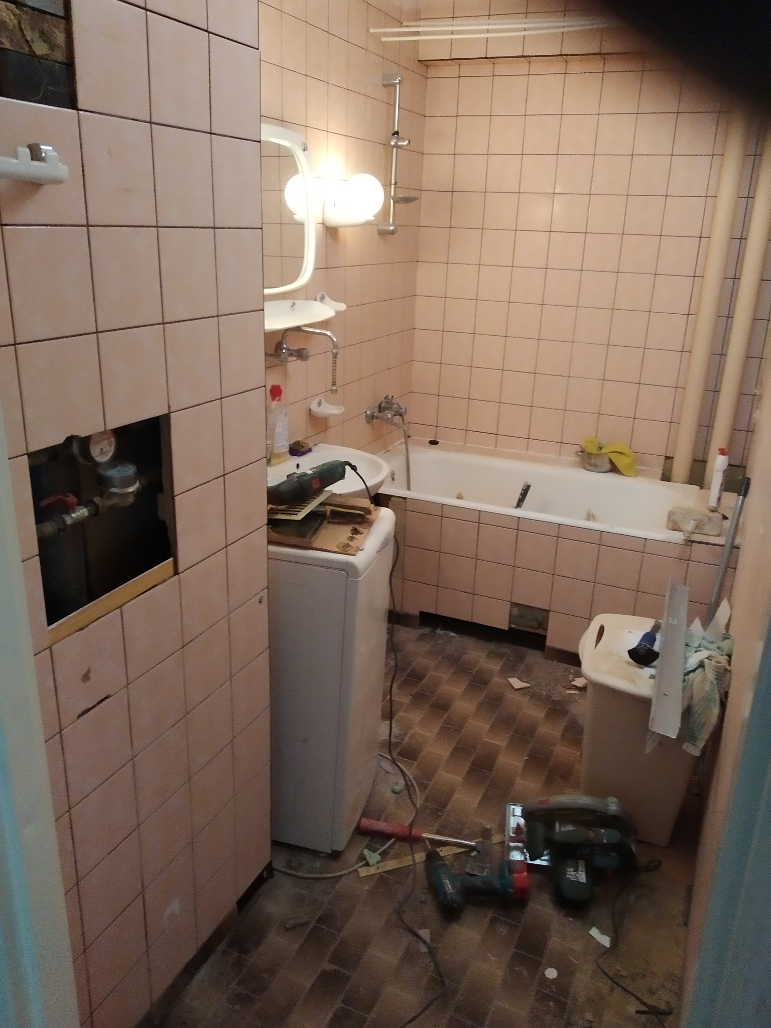 Łazienka w trakcie remontu: częściowo zdjęte kafelki, odsłonięte rury, pralka, narzędzia na podłodze, wanna z żółtą szmatką.