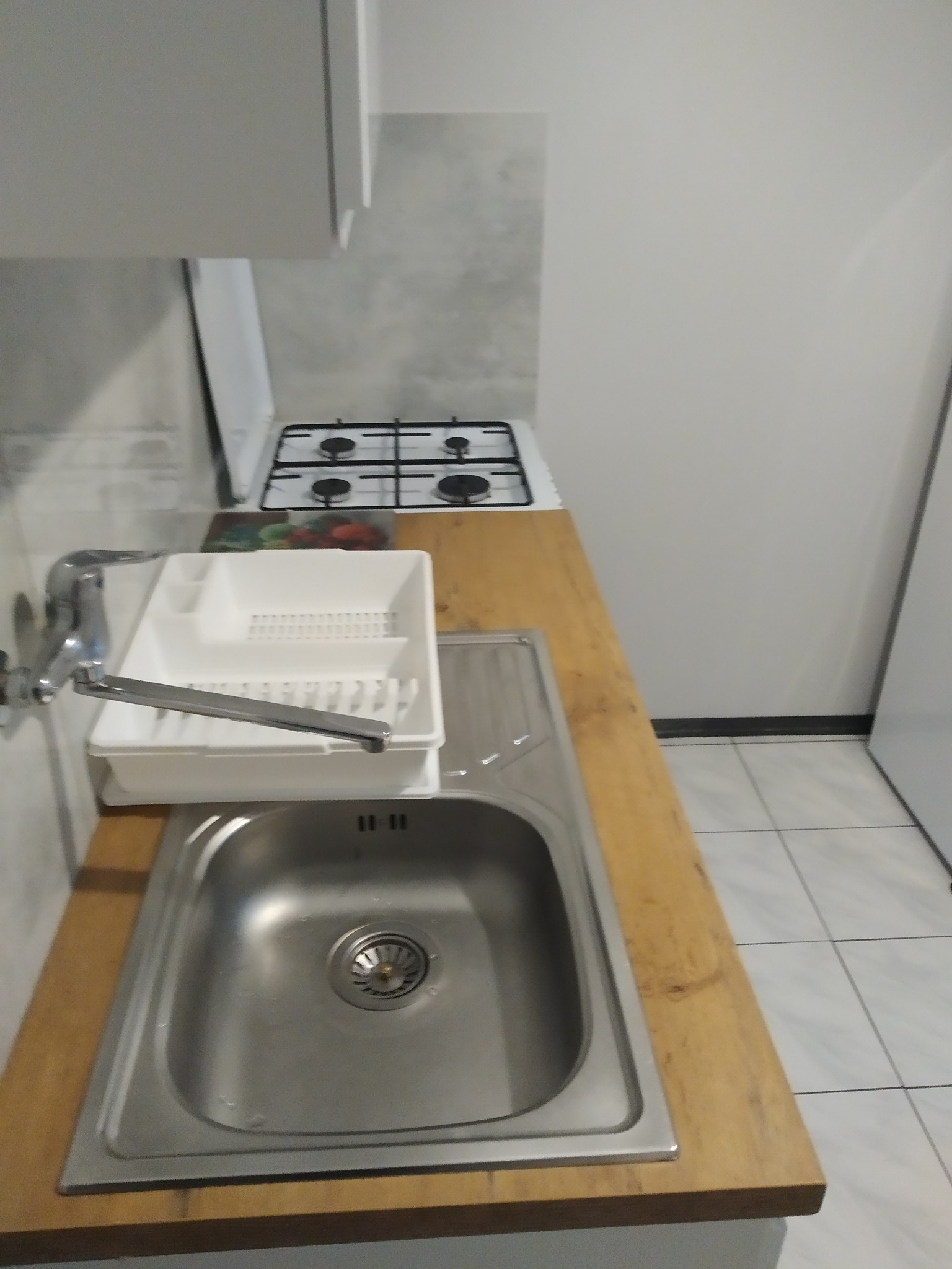 Kątowe ujęcie zlewu ze stali nierdzewnej z baterią i ociekaczem na drewnianym blacie, obok kuchenka gazowa w jasnej kuchni z płytkami podłogowymi.