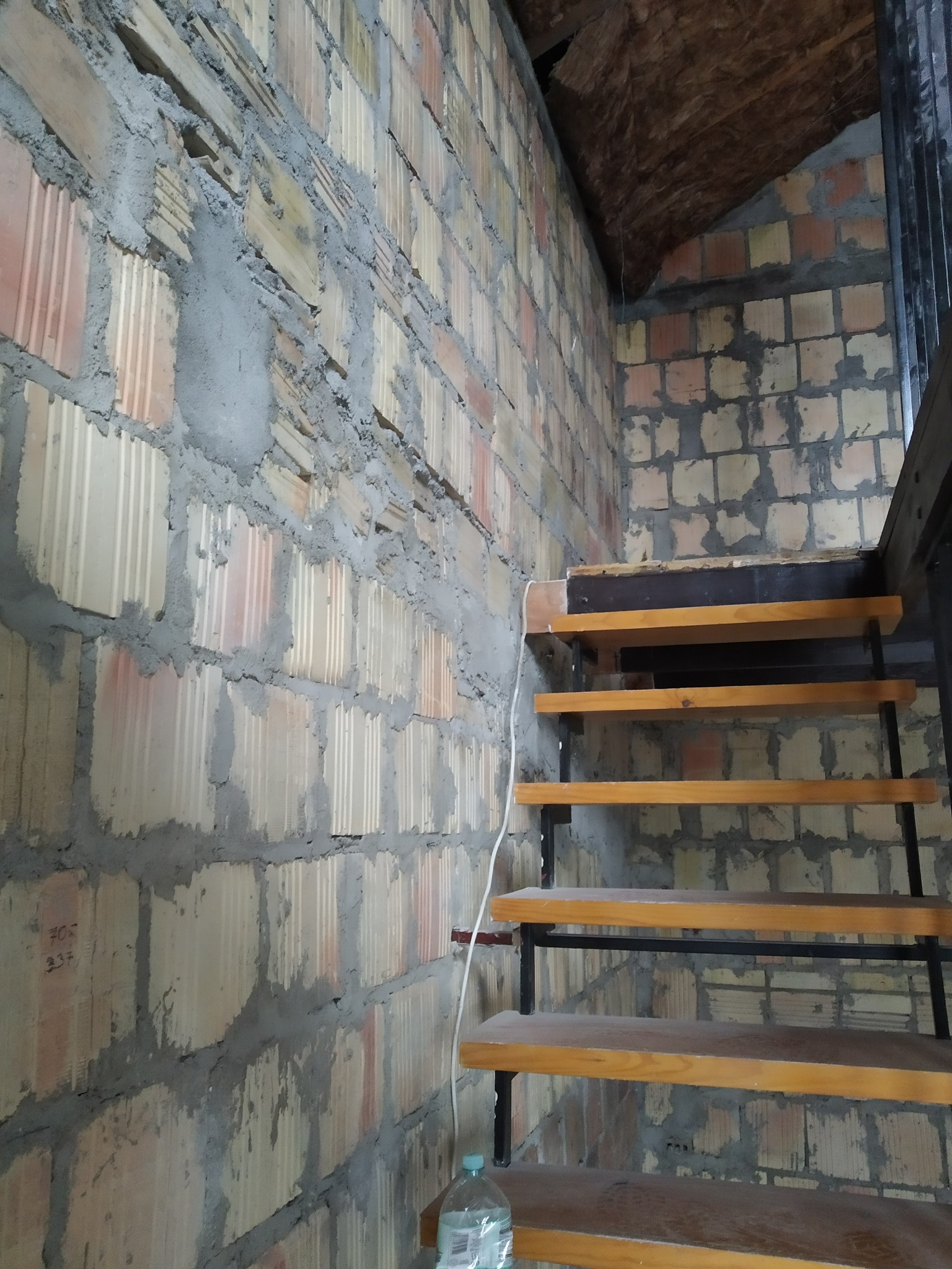 Surowa ściana z bloczków z widocznymi spoinami i fragmentem ocieplonego poddasza, obok schody z drewnianymi stopniami i metalową konstrukcją.